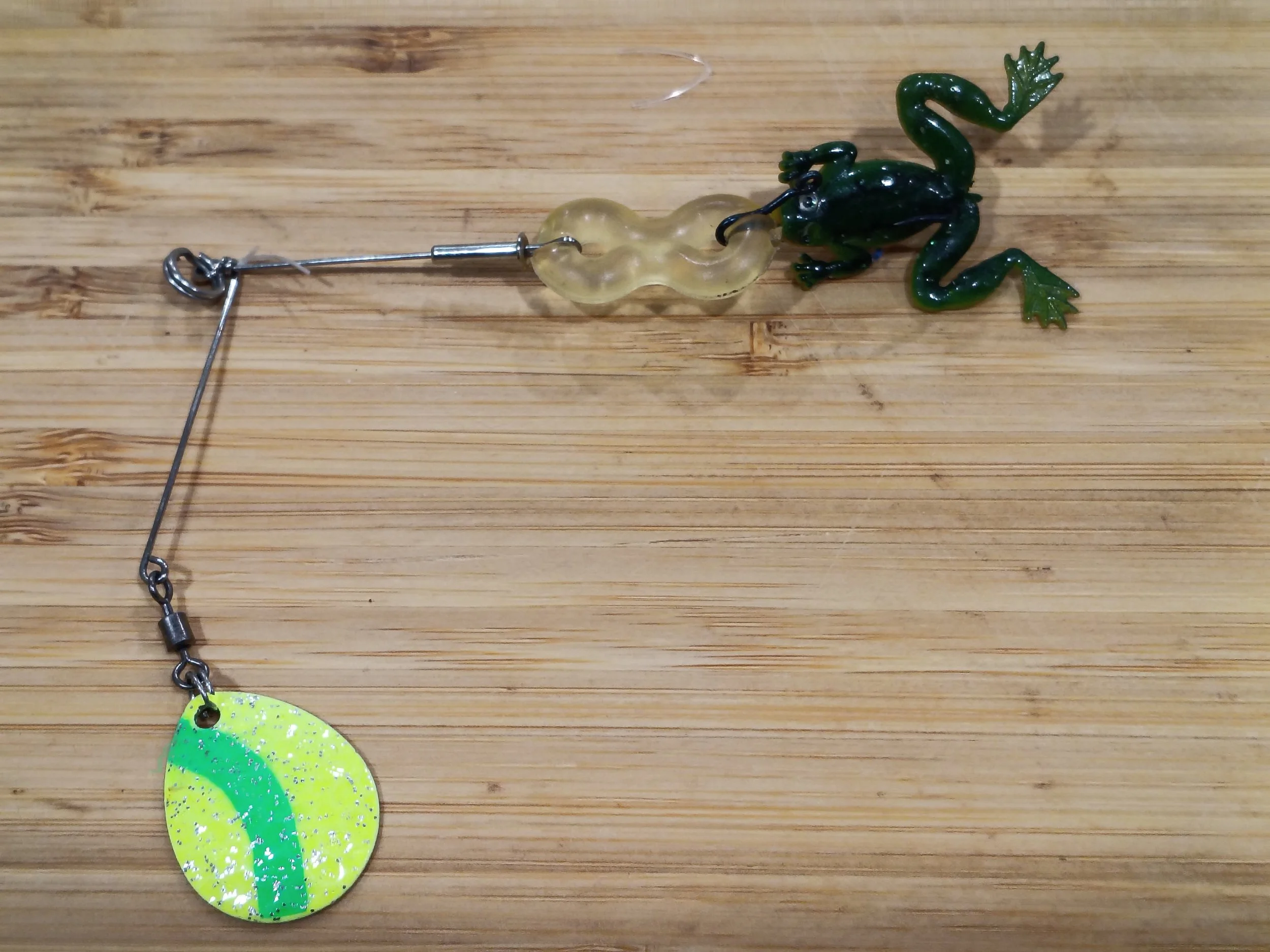 A fishing lure shaped like a frog with a green and black body, a curly tail, and a hook in its mouth, attached to a fishing line on a wooden surface.