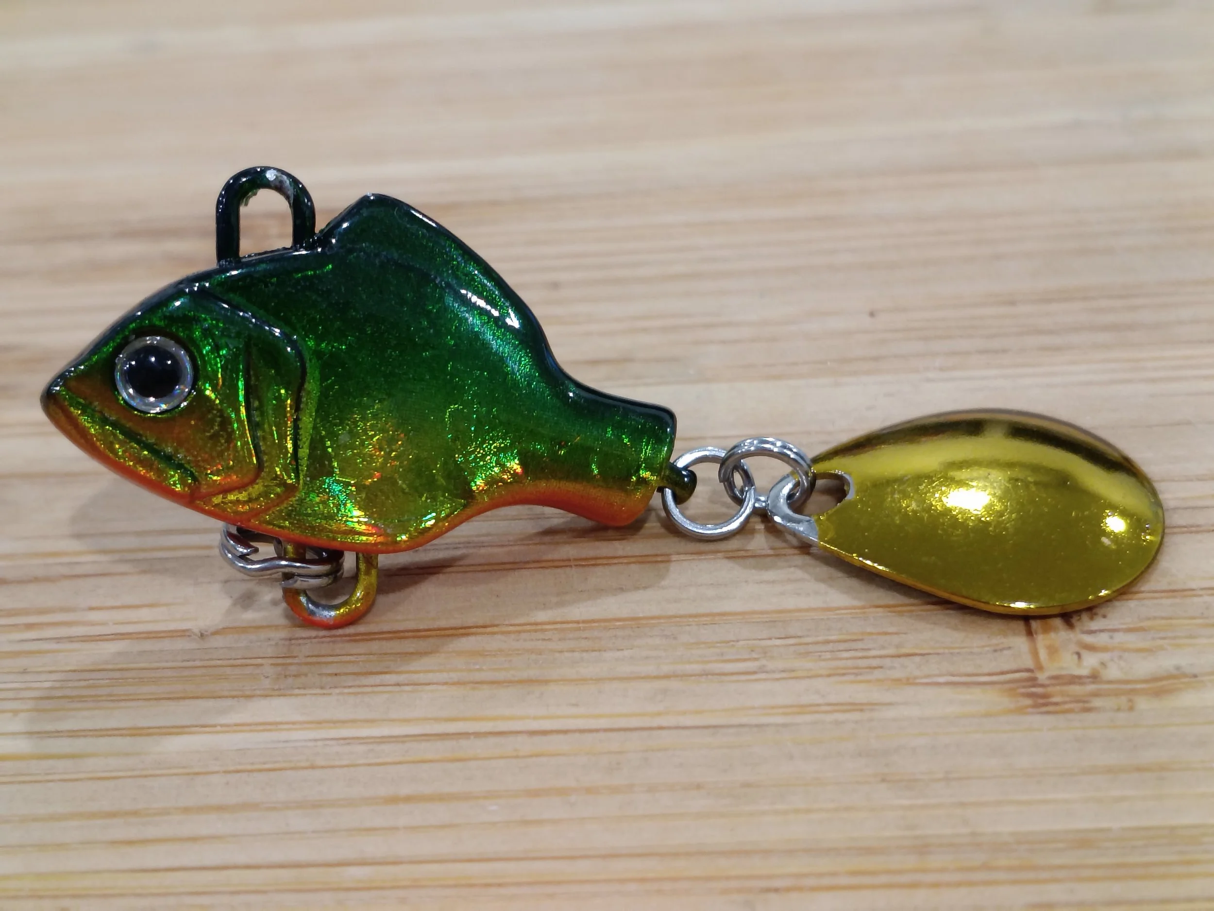 Close-up of a colorful fishing lure shaped like a fish with green, orange, and black reflective hues, attached to a small metal swivel, lying on a wooden surface.