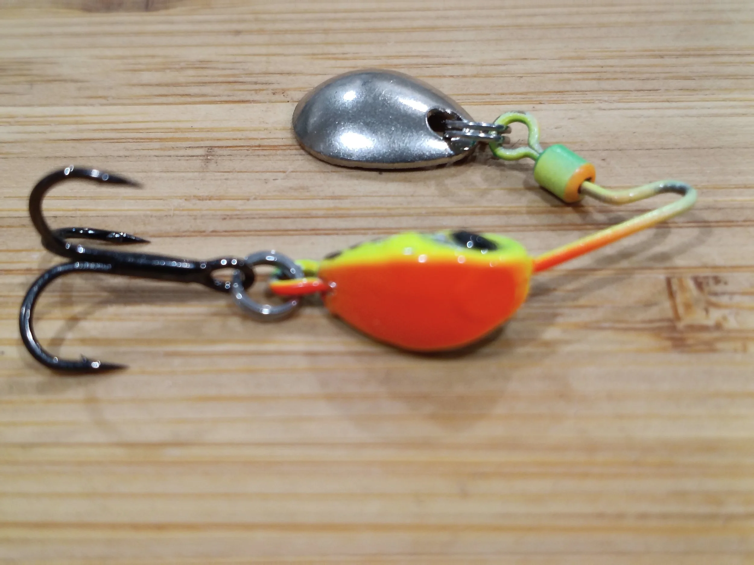 Close-up of fishing tackle, including a metal spoon lure with a hook and a colorful artificial fish bait on a wooden surface.