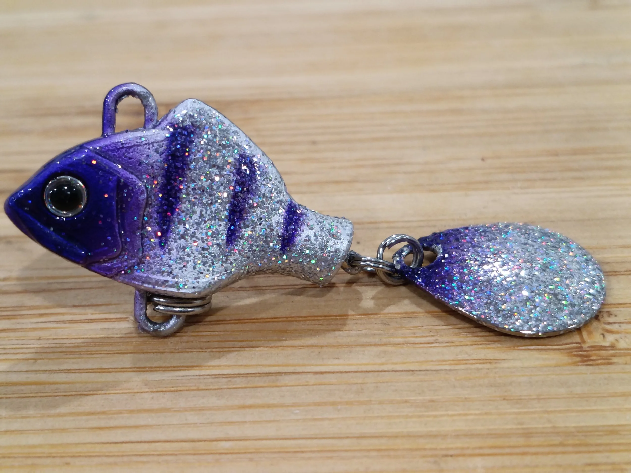 A glittery purple fish-shaped object with a large eye, a hook attached, and sparkles covering its body, resting on a wooden surface.
