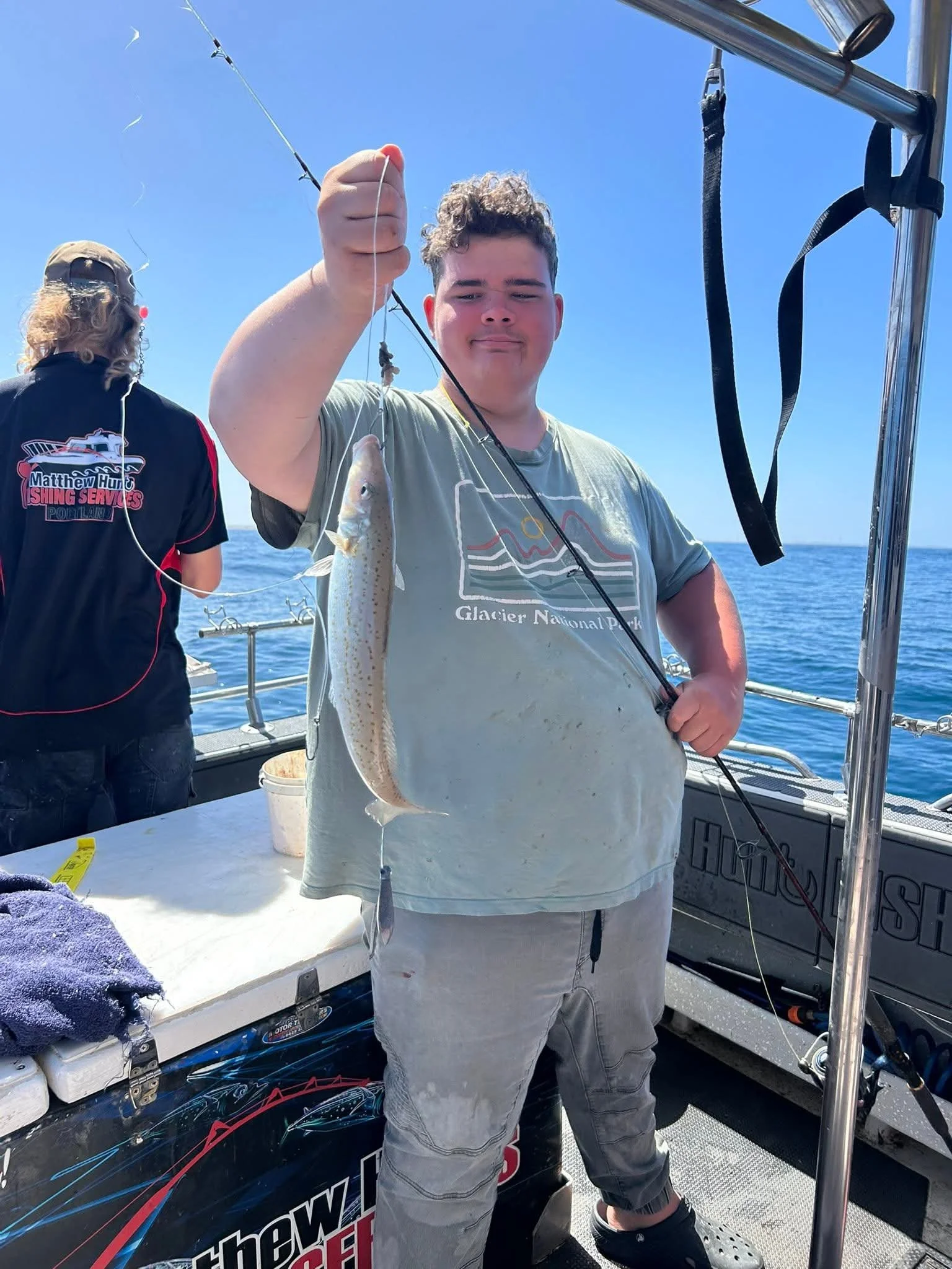 Young man holding a fish on a boat with another person in the background fishing. Clear blue sky and ocean in the background.