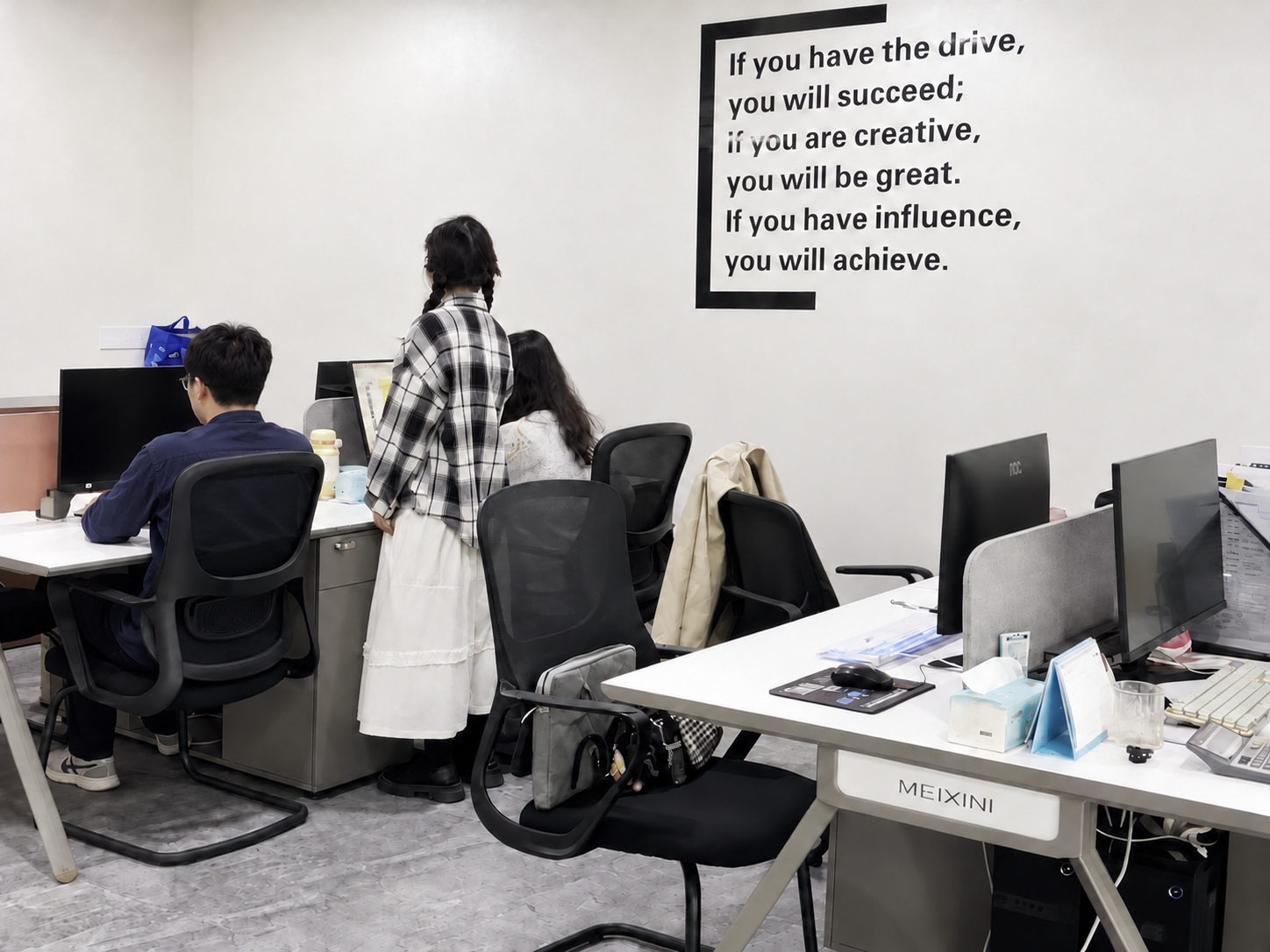 Office workspace with three people working at desks equipped with computers and office supplies. A woman stands talking to two seated colleagues. There is a large motivational poster on the wall with text about success, creativity, influence, and achievement.