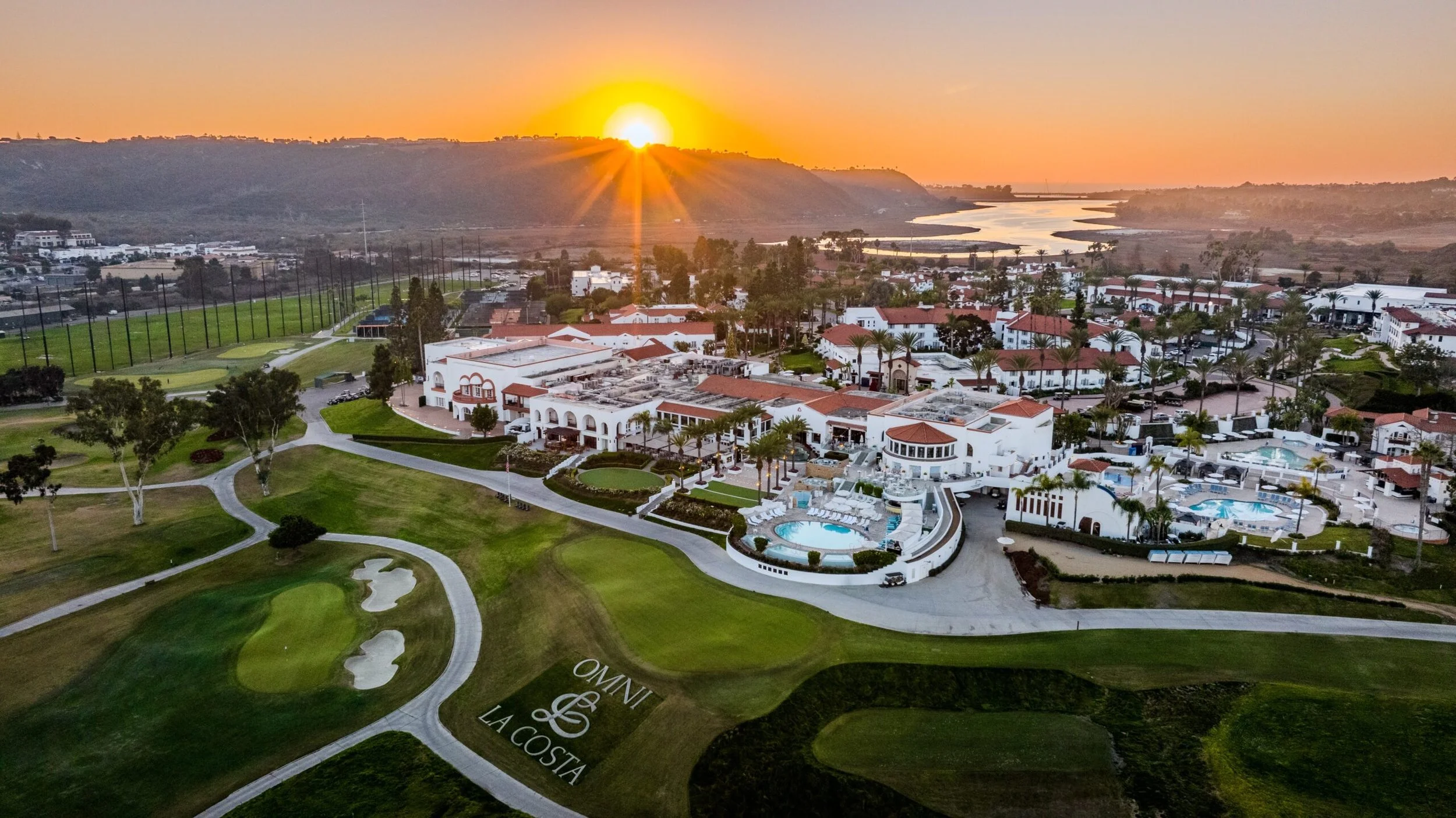Sunset over a golf course and luxury resort with swimming pools, palm trees, and white buildings.