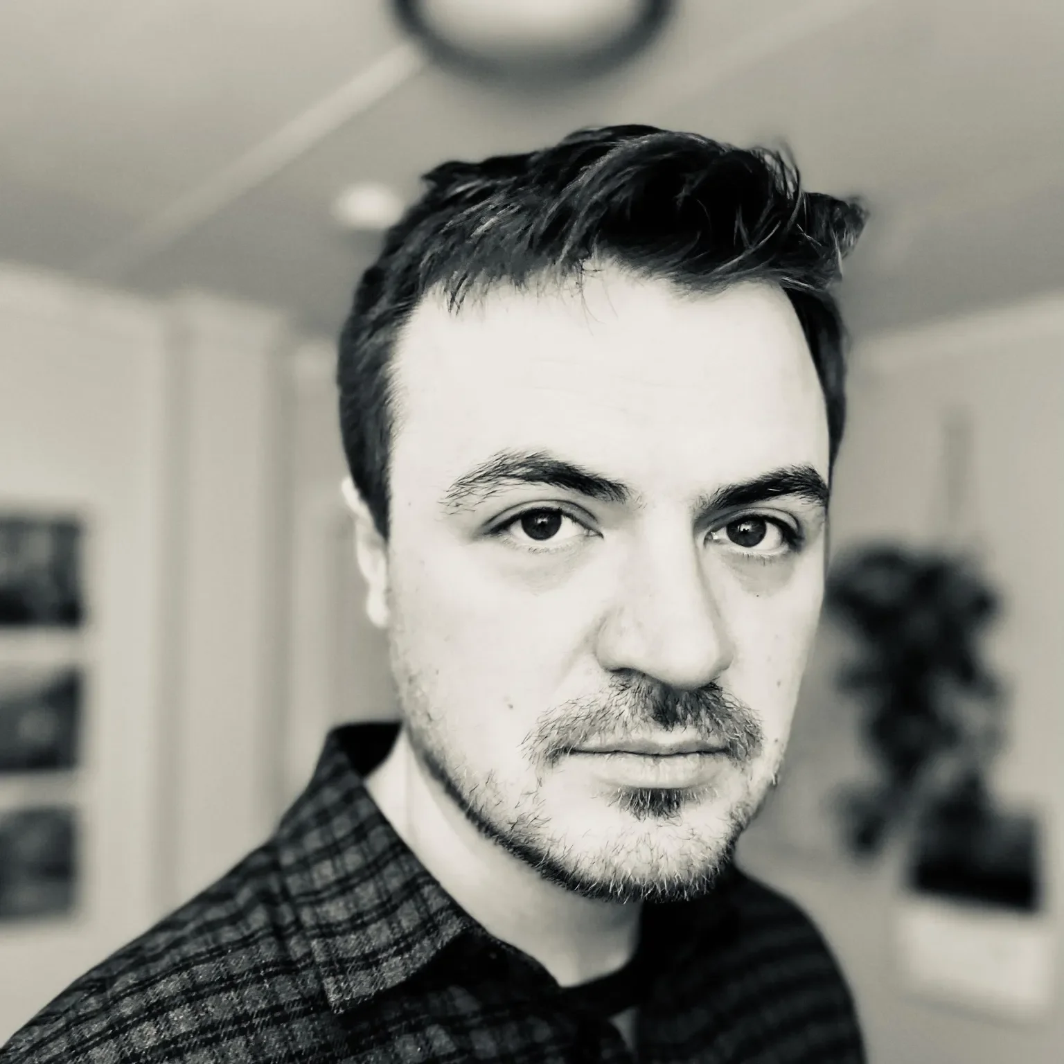 Et svart-hvitt bilde av en mann med mørkt, stygt hår og skjegg, iført en sjekket skjorte, som ser direkte inn i kameraet.