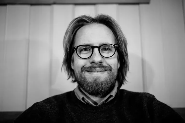 Et svart-hvitt portrett av en mann med briller og skjegg, som smiler litt, foran en vegg med panel.