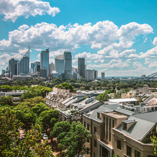 View of a city skyline with tall skyscrapers and a partly cloudy sky, with residential buildings and green trees in the foreground.