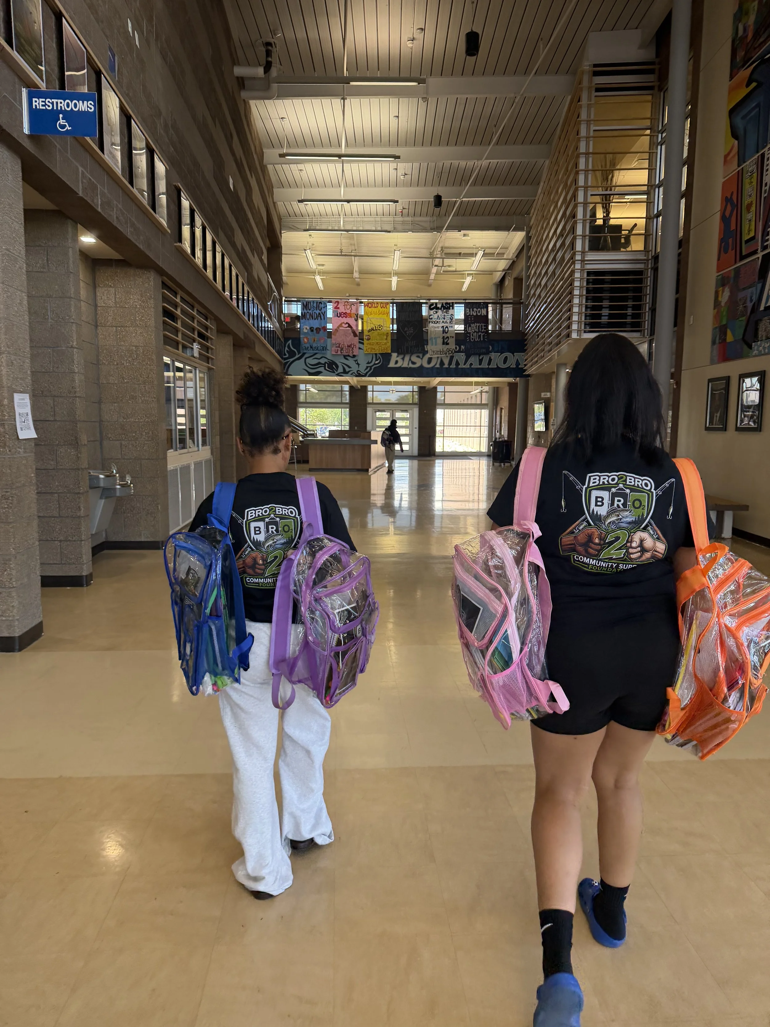 Two young girls walking in a school hall, carrying clear backpacks filled with school supplies. The girl on the right has dark hair and is wearing a black t-shirt, while the girl on the left has curly hair tied up and is dressed in white pants and a dark top. There are colorful banners and artwork hanging in the background.