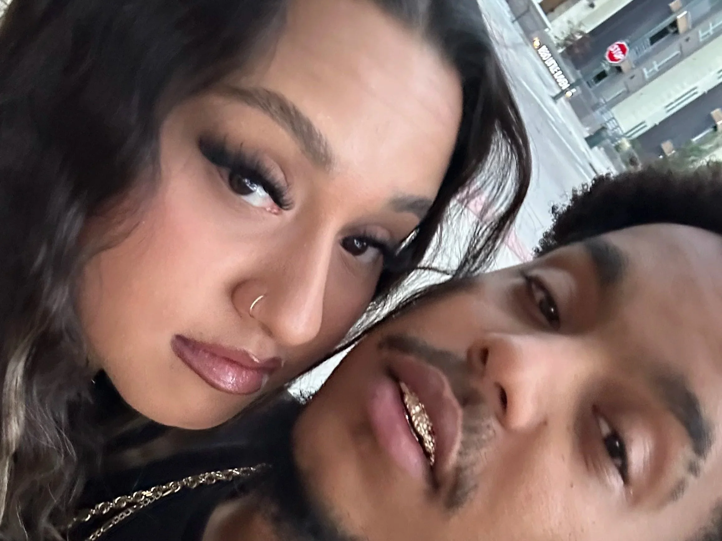 Close-up of two women, one with curly dark hair and makeup, and the other with wearing grills, in an outdoor urban setting.