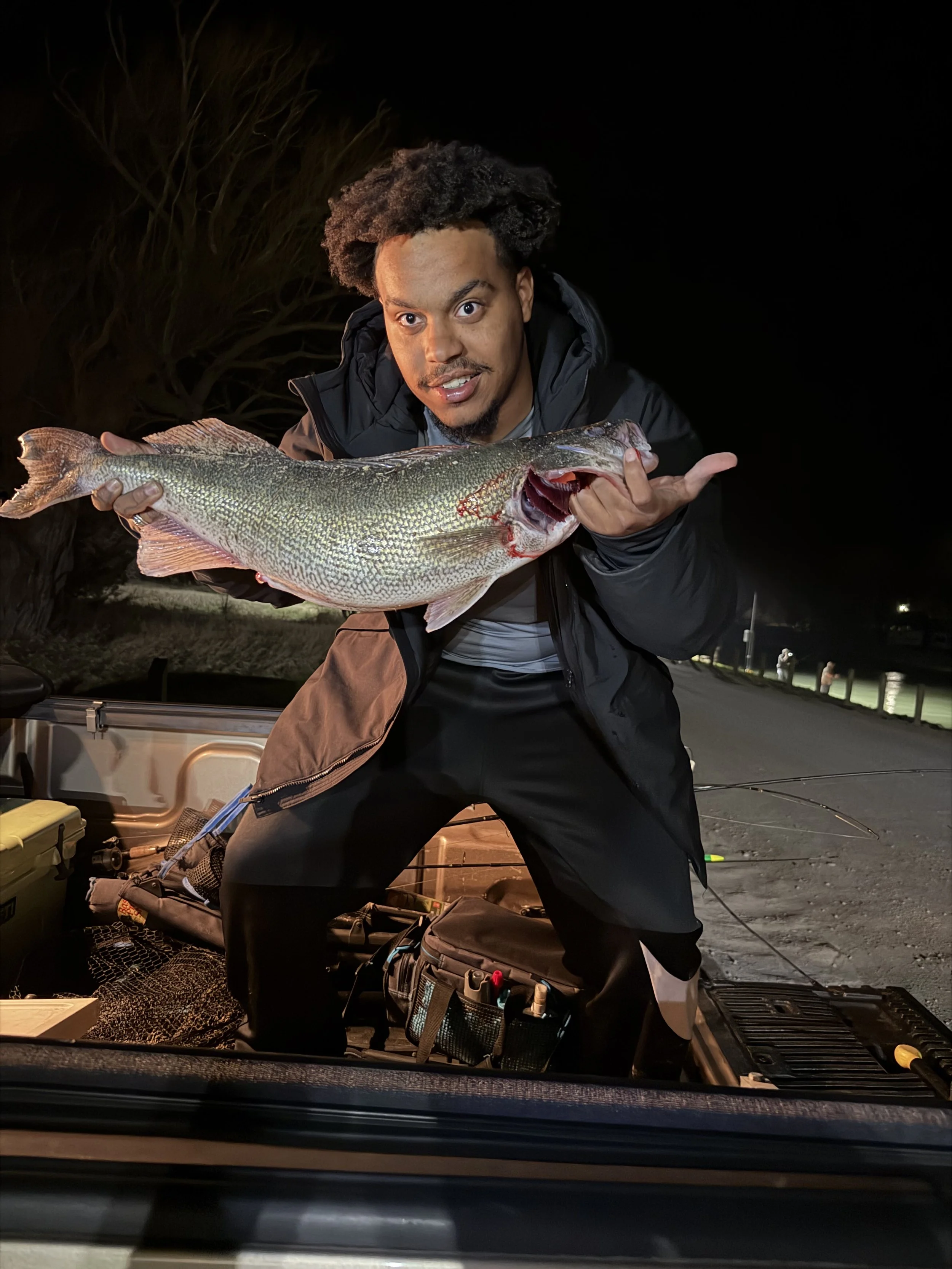 A man holding a large fish, standing at night with fishing gear and supplies visible in the background.