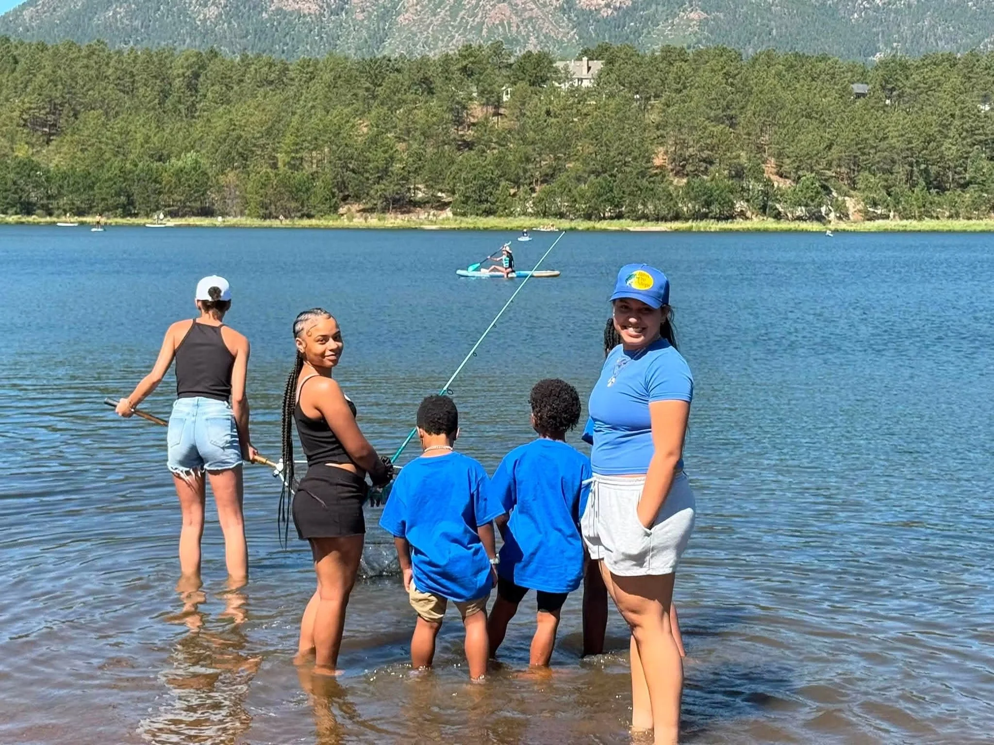 People standing in shallow water at a lake, some wearing blue shirts and others in black and gray, with trees and mountains in the background. One person is paddleboarding in the distance.