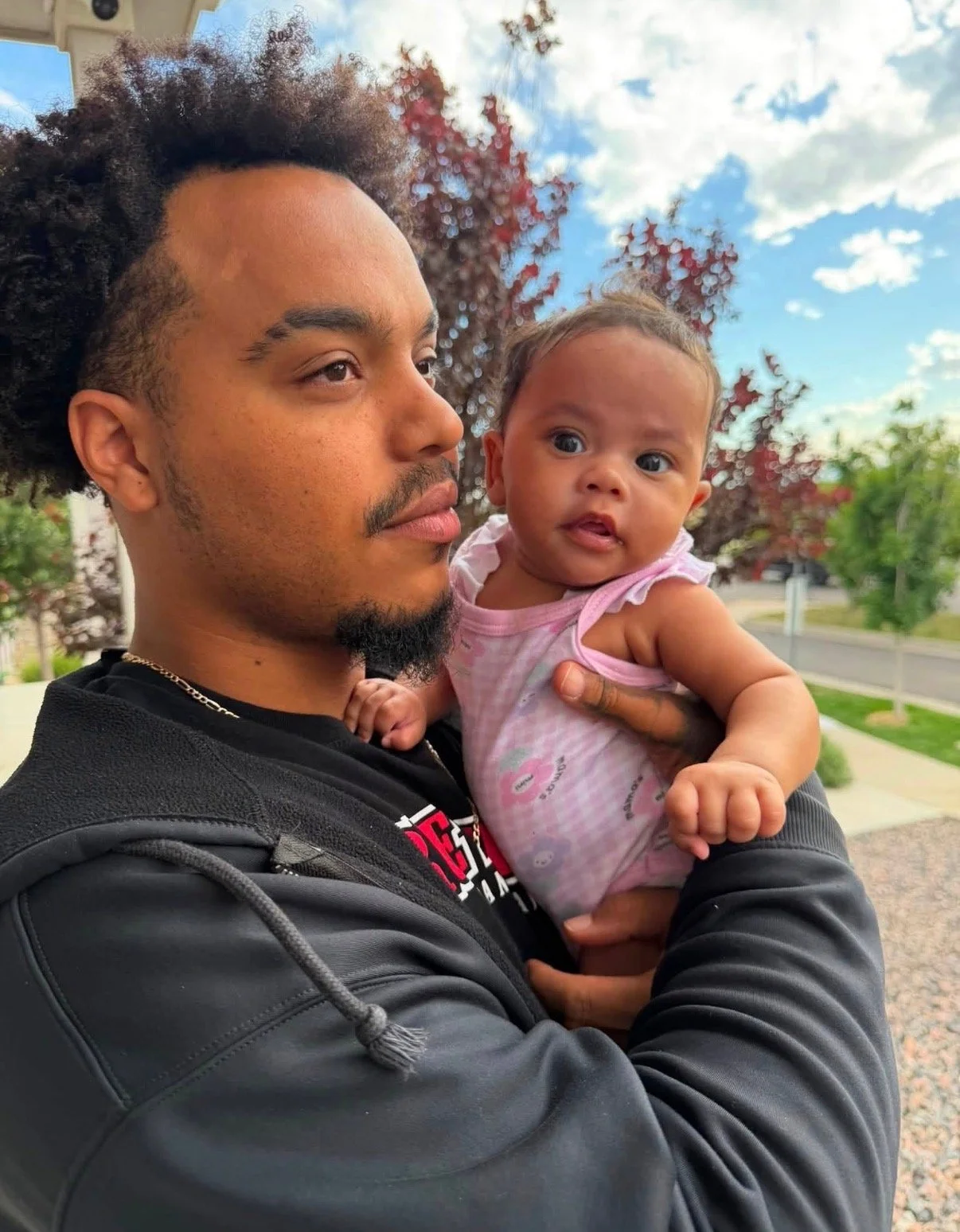 A man holding a young child outdoors on a sunny day, with a background of trees and a partly cloudy sky.