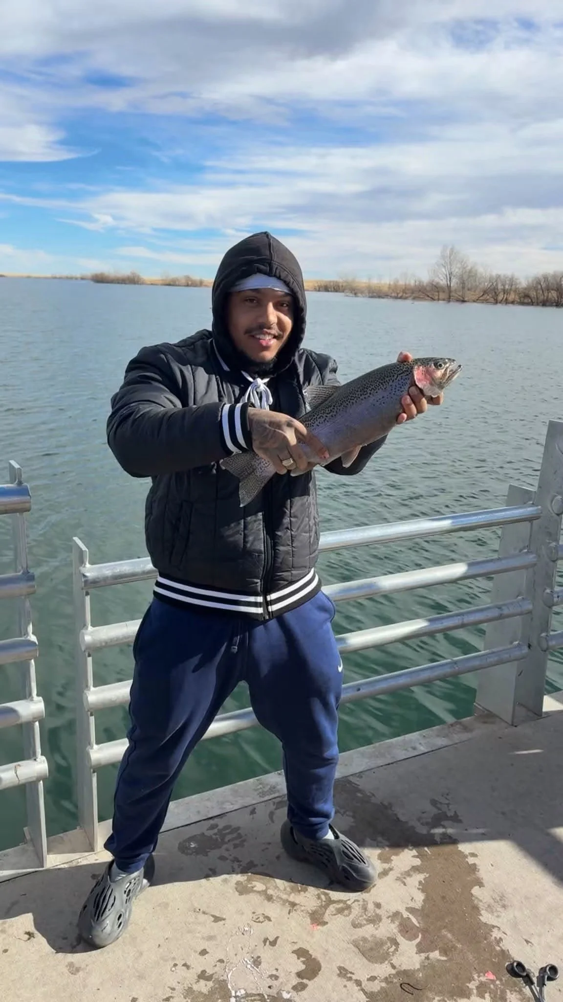 Man holding a large fish on a boat pier with water and trees in the background.