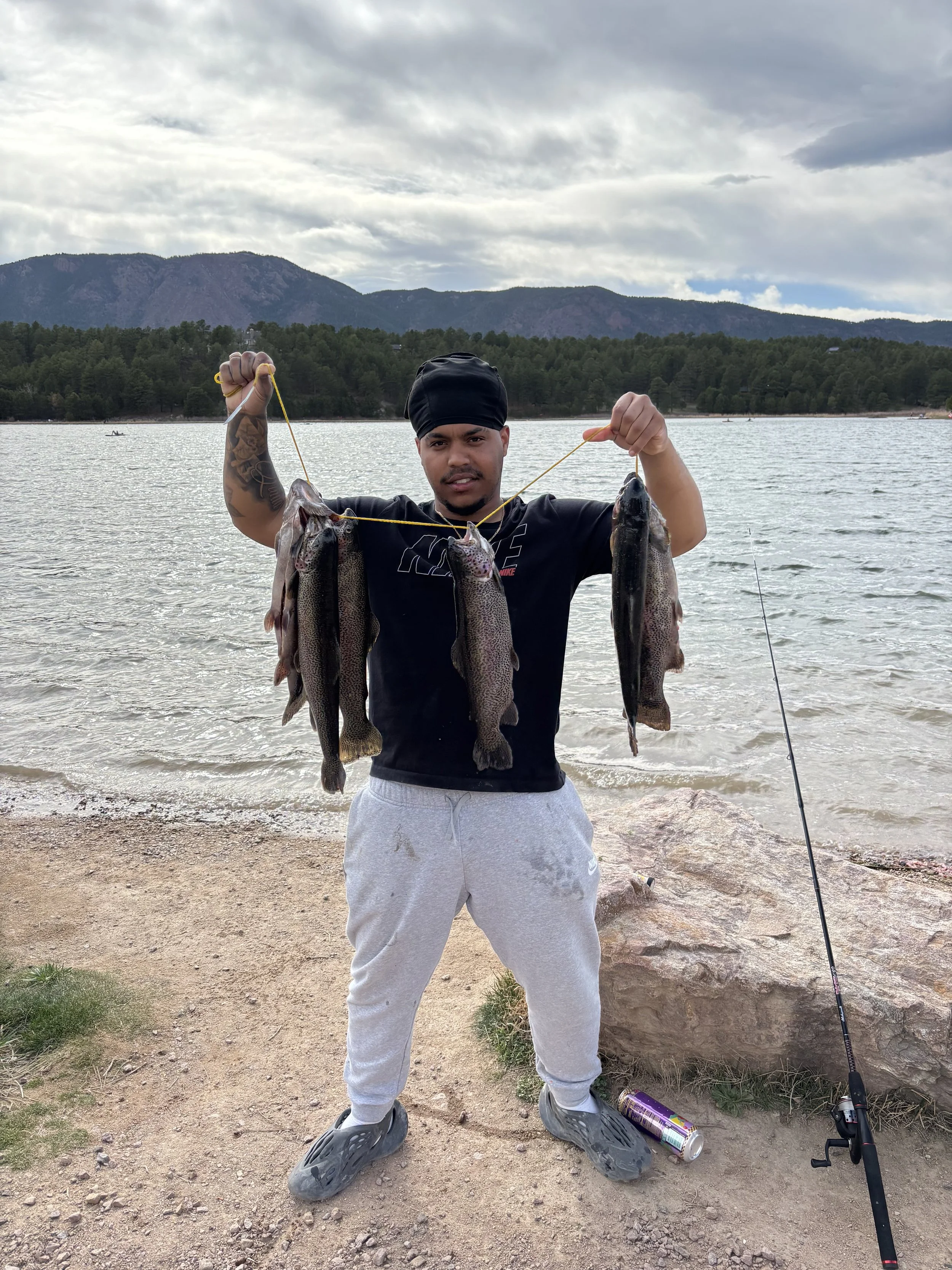 A man standing by the water holding four large fish with mountains and cloudy sky in background