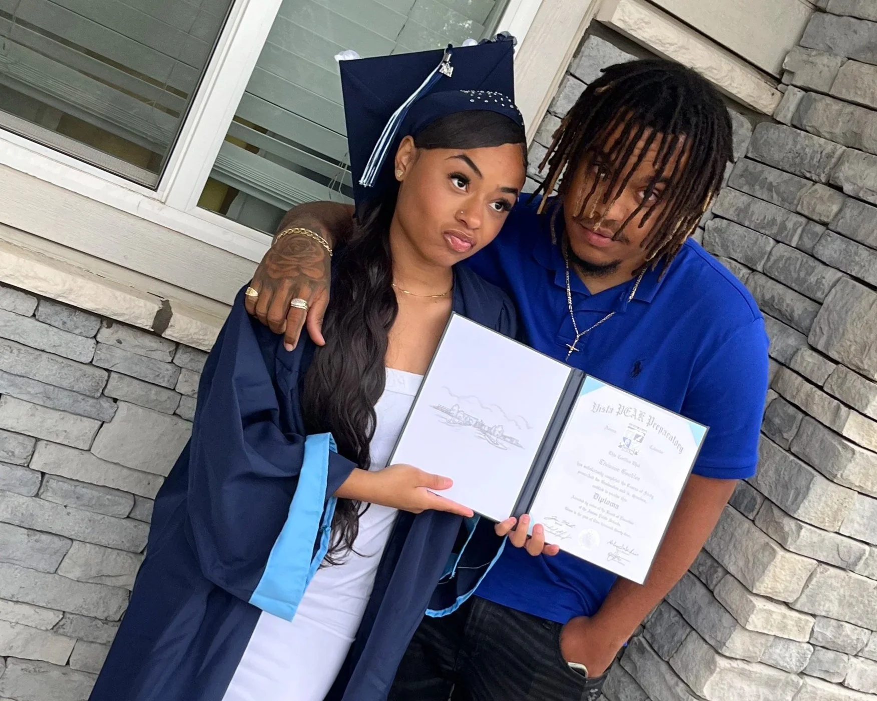 A young woman in a graduation cap and gown holding her diploma, standing next to a young man who has his arm around her shoulders. They are outside in front of a stone and siding wall.