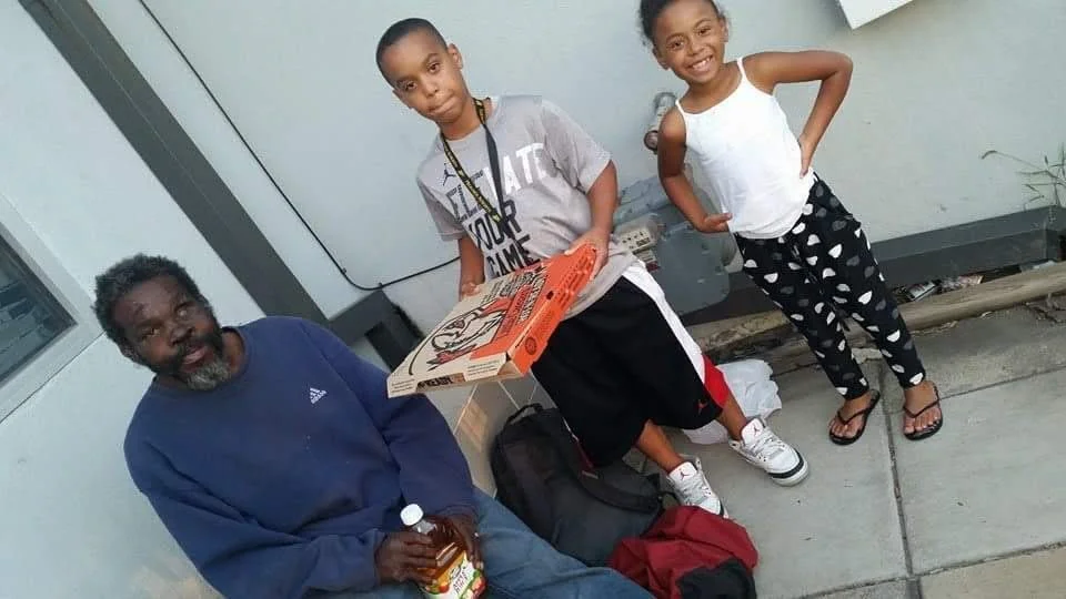 Three children standing and sitting outside near a building with a man who is sitting and holding a bottle, with backpacks and a pizza box on the ground.