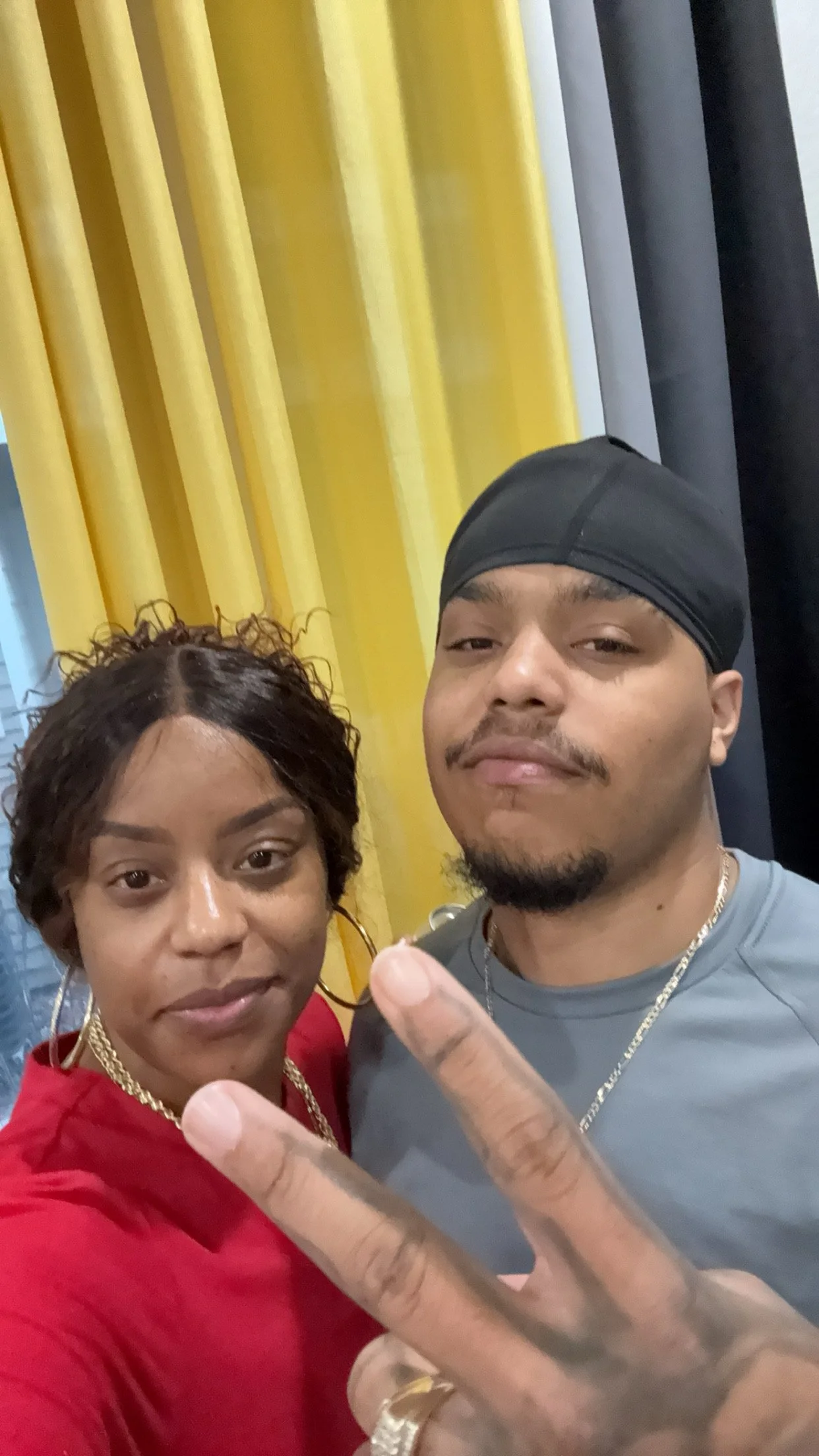 A woman with short curly hair and a man with a beard and a black durag posing for a selfie in front of yellow and gray curtains. The woman is wearing large hoop earrings, a gold necklace, and a red top. The man is wearing a gray shirt, a gold chain, and is making a peace sign with his fingers.