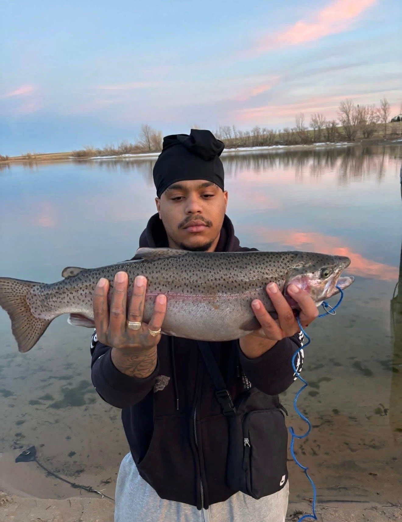 A man holding a large fish near a calm river at sunset with pink and blue sky reflected in the water.