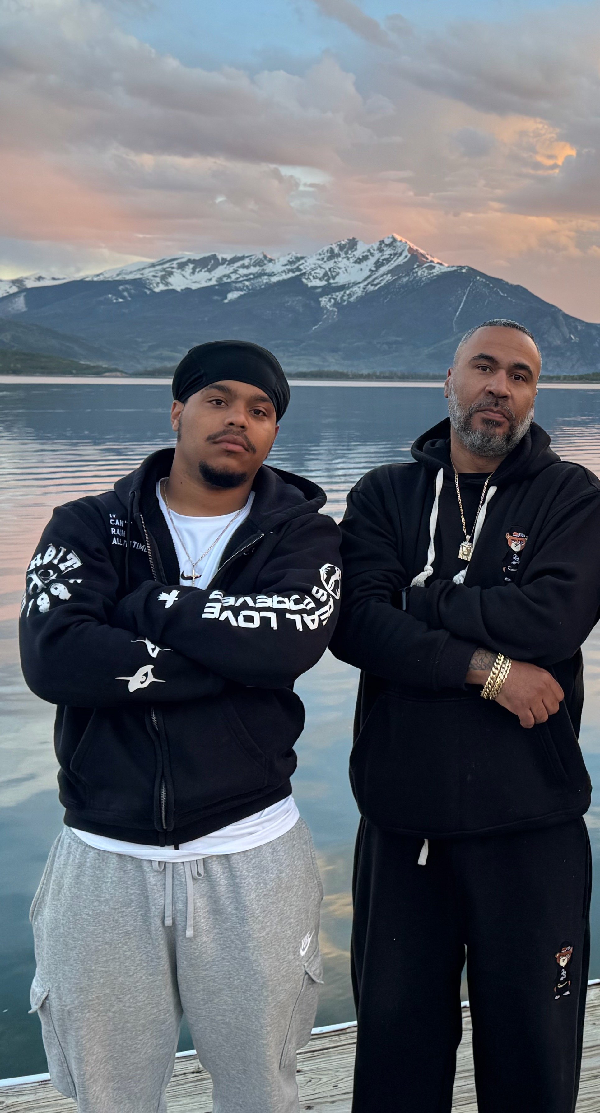 Two men standing in front of a lake with snow-capped mountains and a cloudy sky in the background.