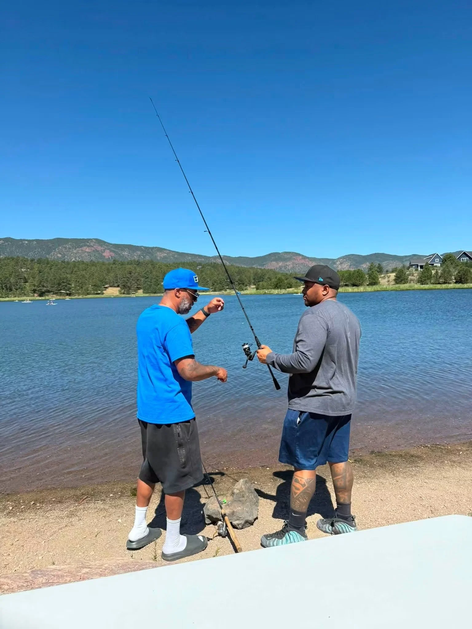 Two men are fishing at the edge of a lake with forest and mountains in the background. They are standing on a sandy shore, one wearing a blue cap and shirt, the other in a black cap and gray shirt, both holding fishing rods.