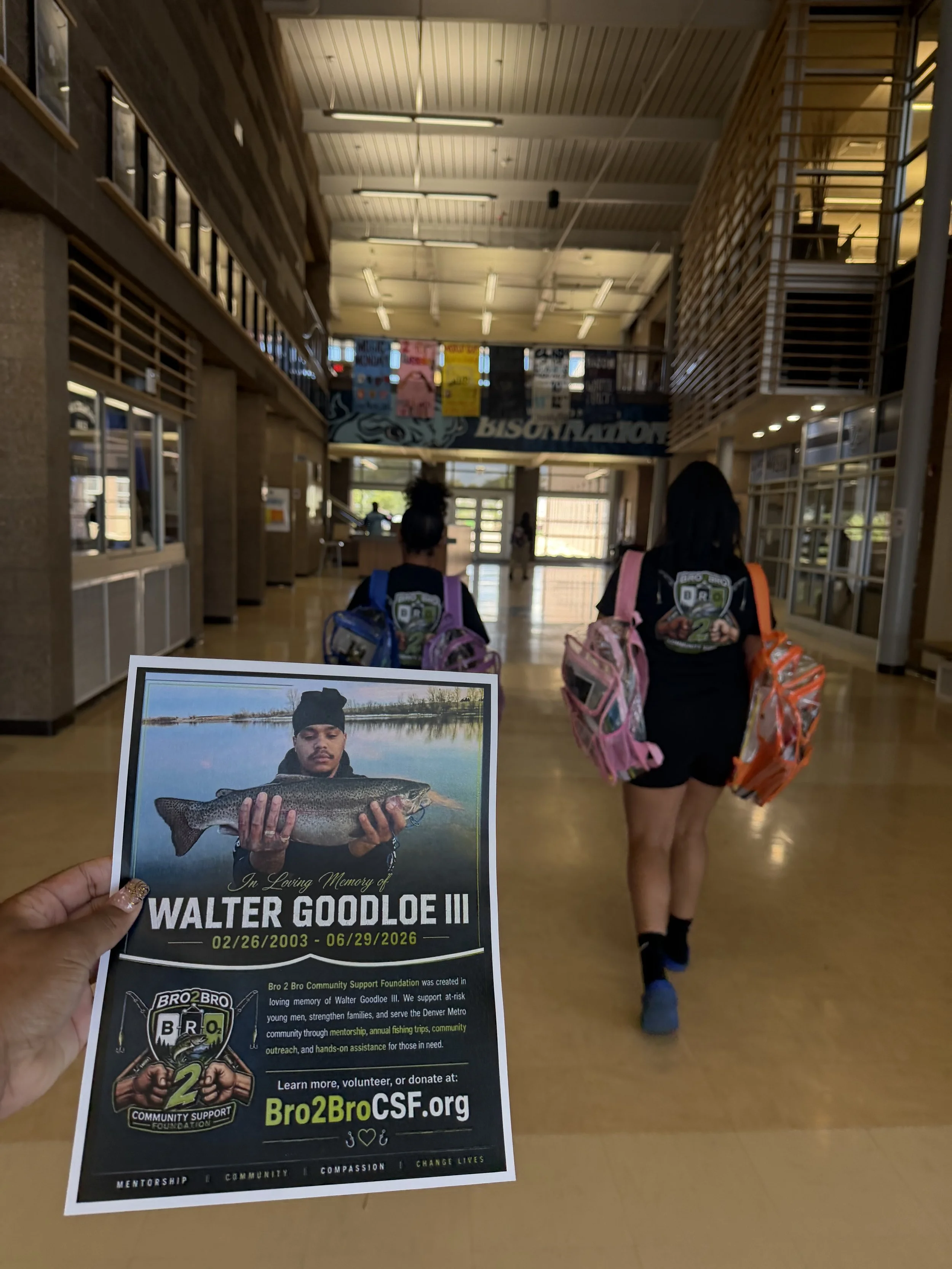 A person holding a memorial poster for Walter Goodloe III, showing a young man holding a fish, with dates of birth and death, inside a building with people walking in the background.