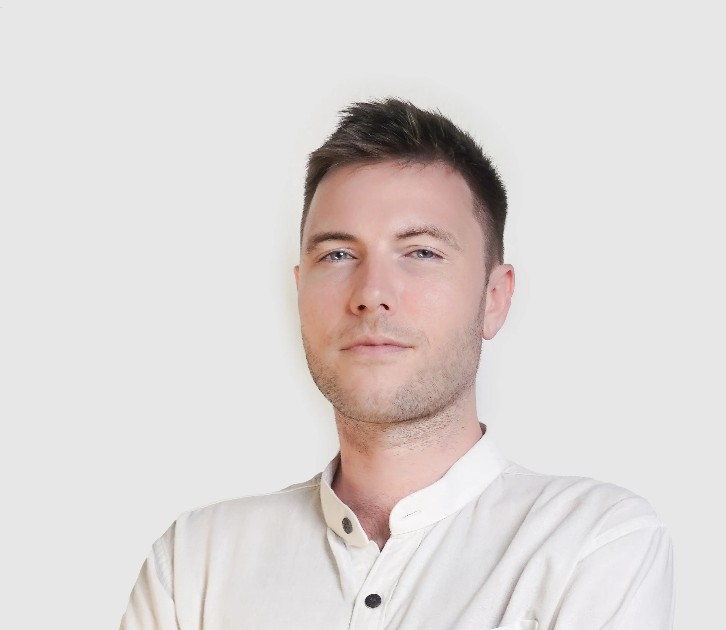 A young man with blue eyes and short, dark, styled hair wearing a white button-up shirt against a plain white background.