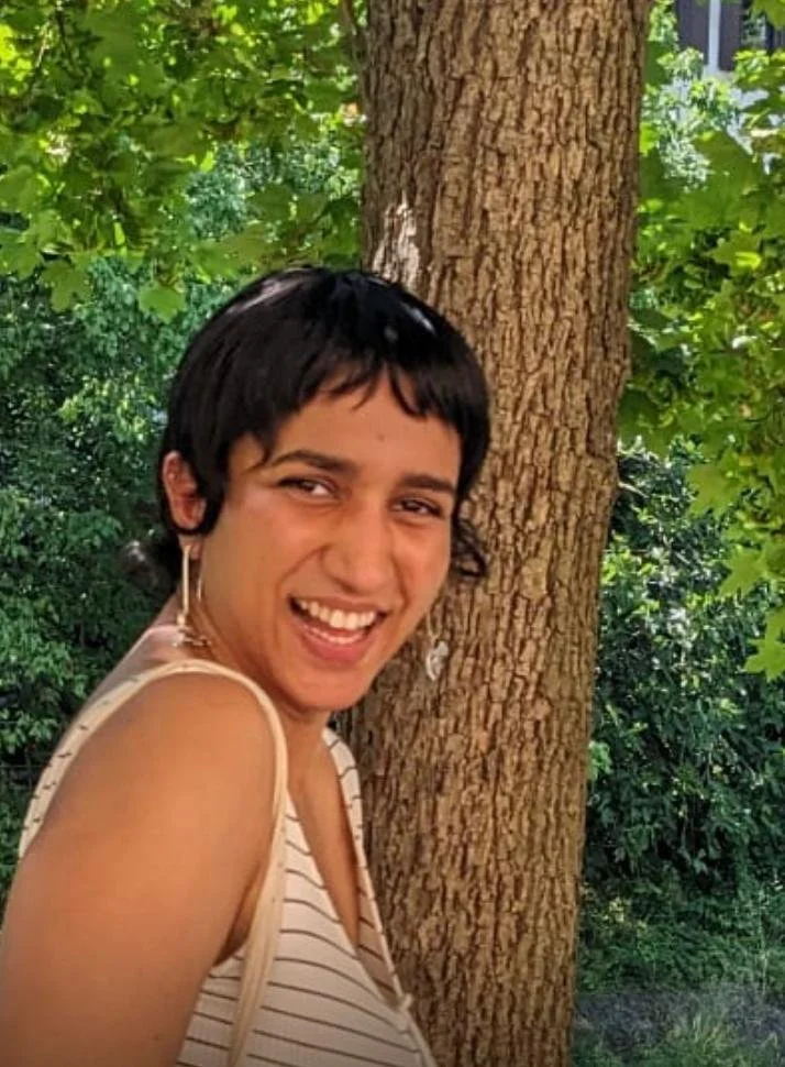 A smiling woman with short dark hair standing next to a tree in a green outdoor area.