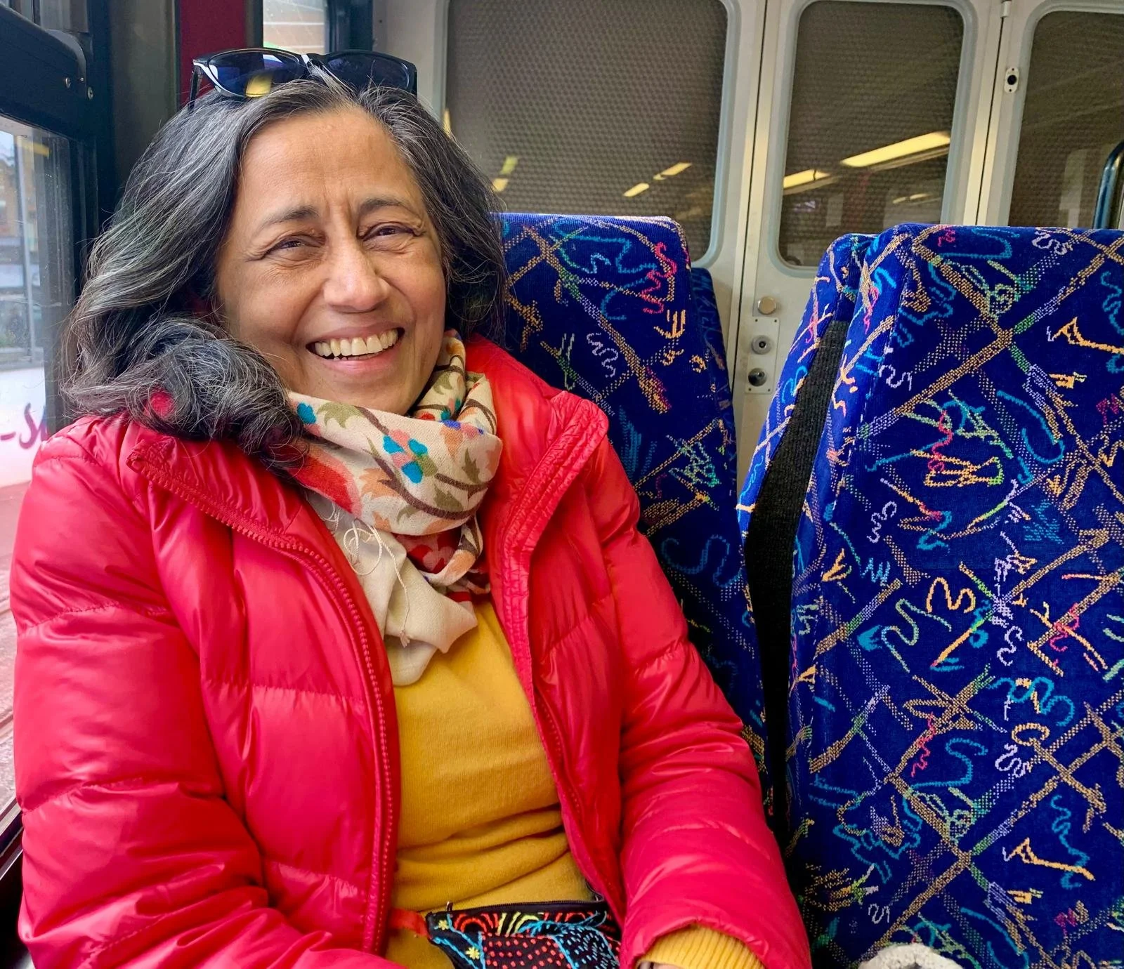A woman sitting on a bus with a bright smile, wearing a red jacket, a yellow shirt, a colorful scarf, and sunglasses on her head, with patterned blue bus seats around her.