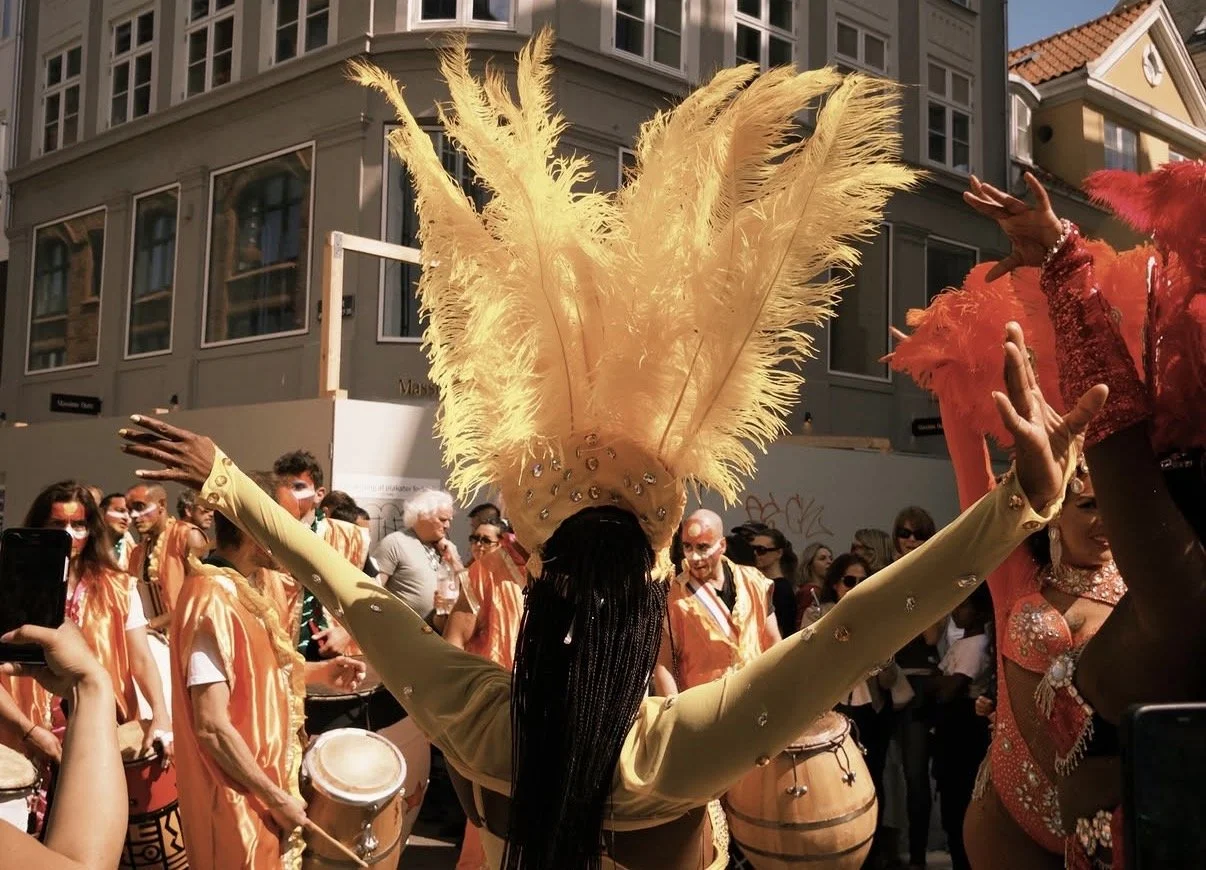 En person med stort gult fjäderhuvud och gul klädsel dansar i en grupp under en parade eller festival, med folk i bakgrunden.