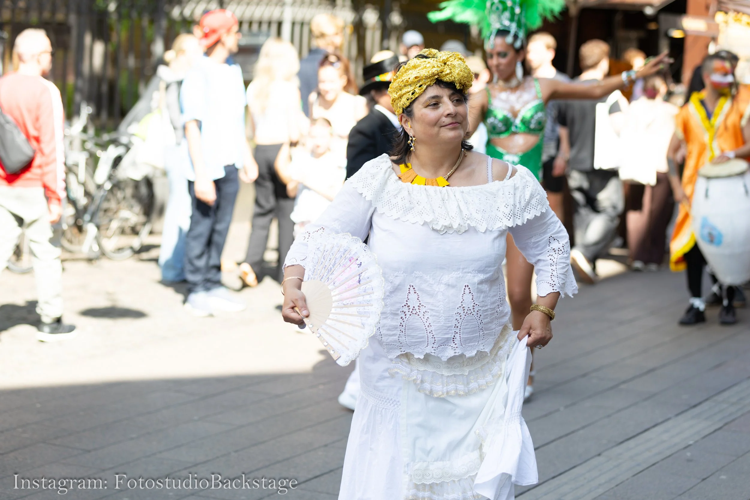 Kvinna i vit klänning med gul huva och handfläkt, deltar i en paradränna under en festival eller karneval, omgiven av andra människor i färgglada eller traditionella kläder.