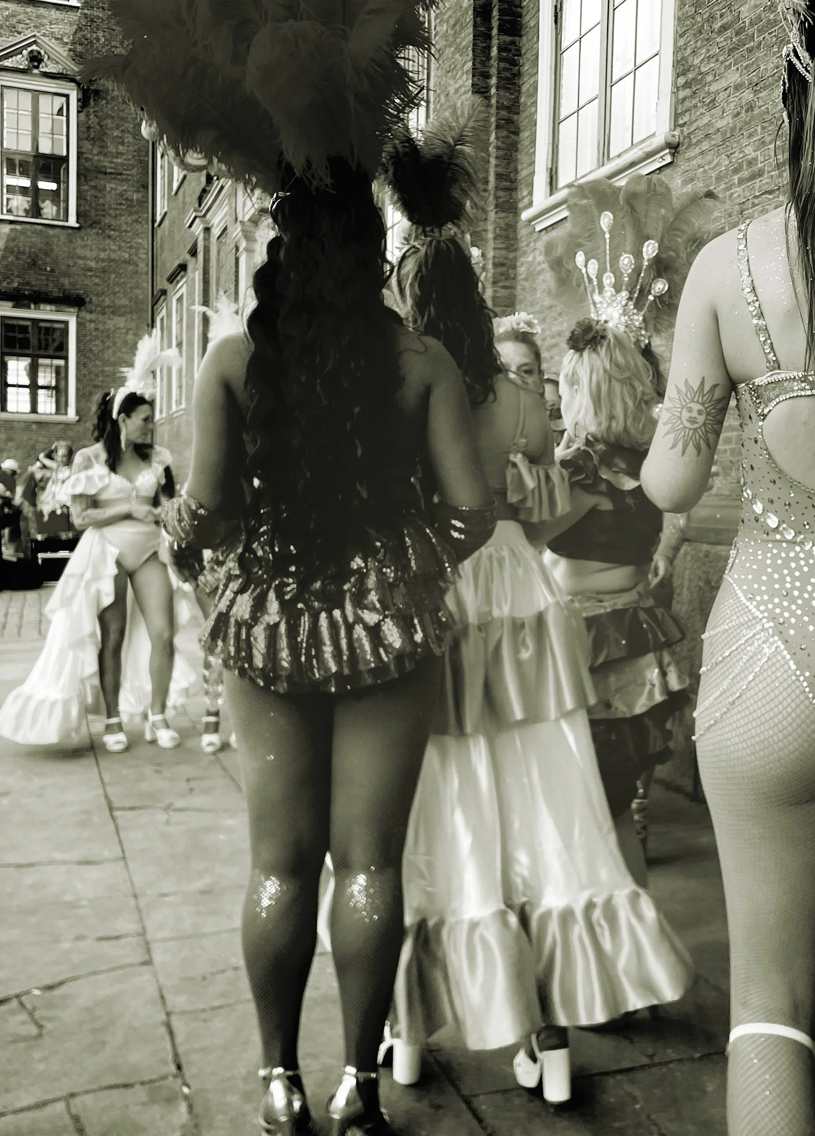 Kvinnor i glamourösa kostymer och festskor på en gata, med en byggnad i bakgrunden, troligen under en festival eller karneval.