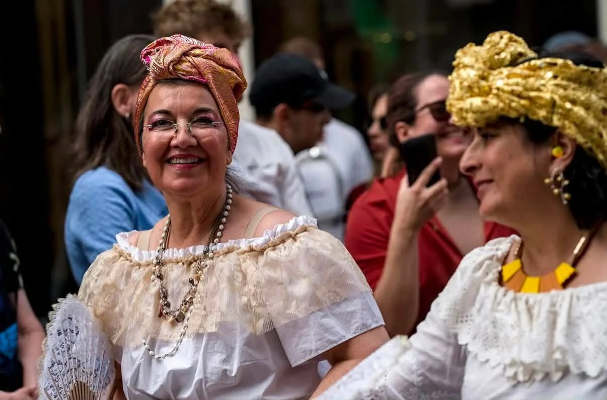 Två kvinnor i traditionella kläder och med huvudbonader, omgivna av människor, på en fest eller festival.