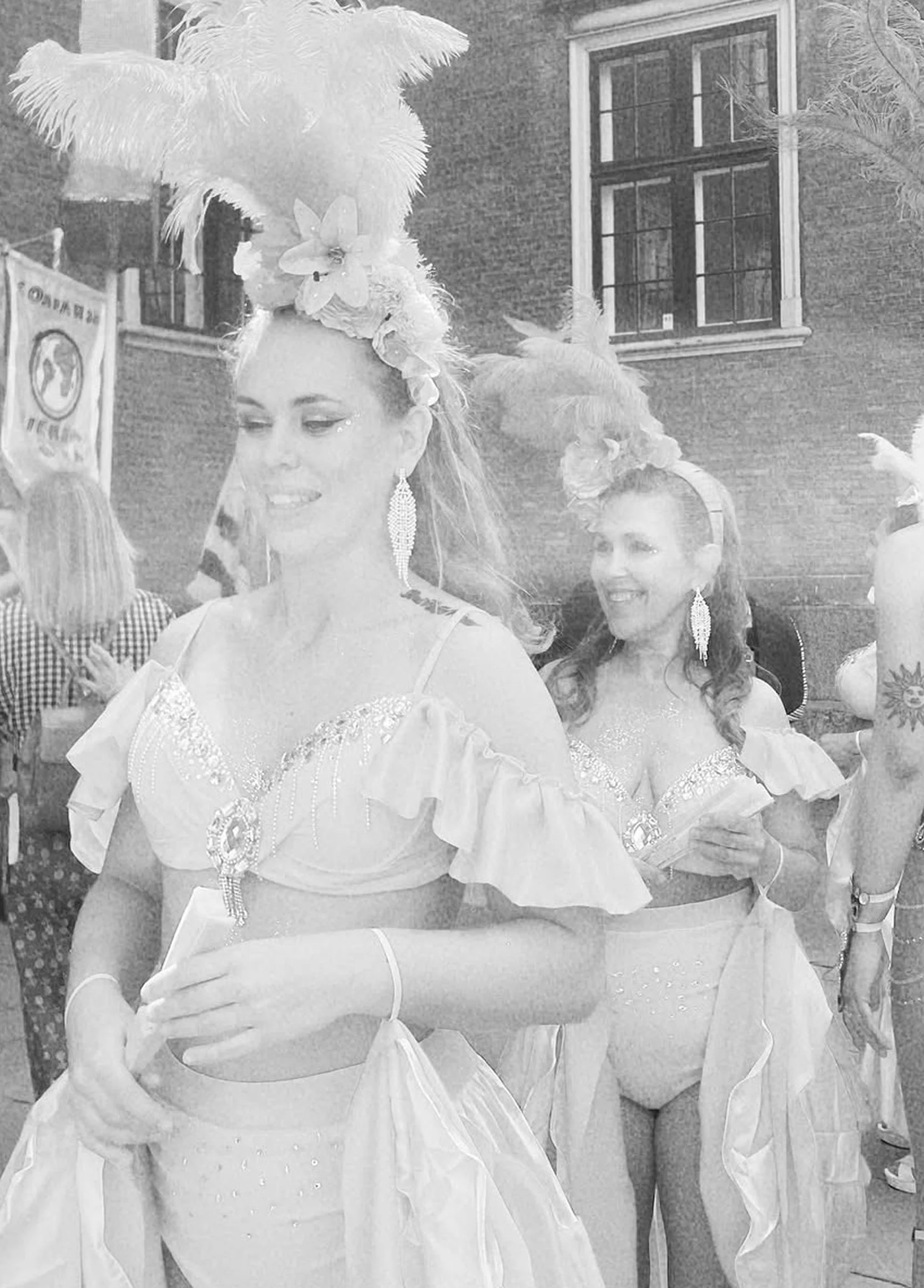 Två kvinnor i karneval dräkt, en av dem har en stor fjäderhatt och diamanter, den andra bär stora örhängen, står utomhus framför ett tygtak och en tegelbyggnad.