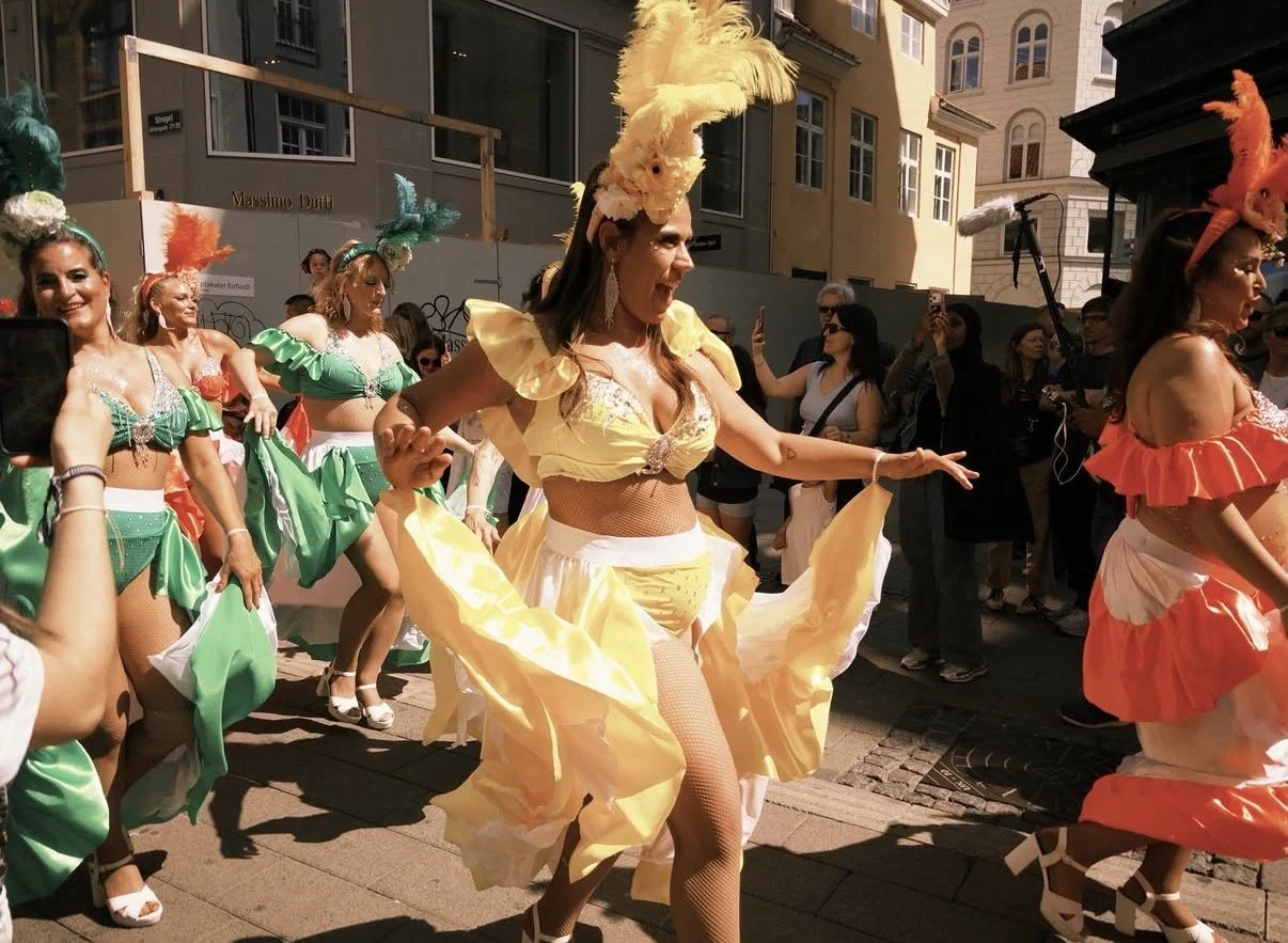 Flera kvinnor i färgglada, extravagant klädsel och fjäderhuvor dansar på en gata under en parad, med en publik som filmar och tar bilder i bakgrunden.
