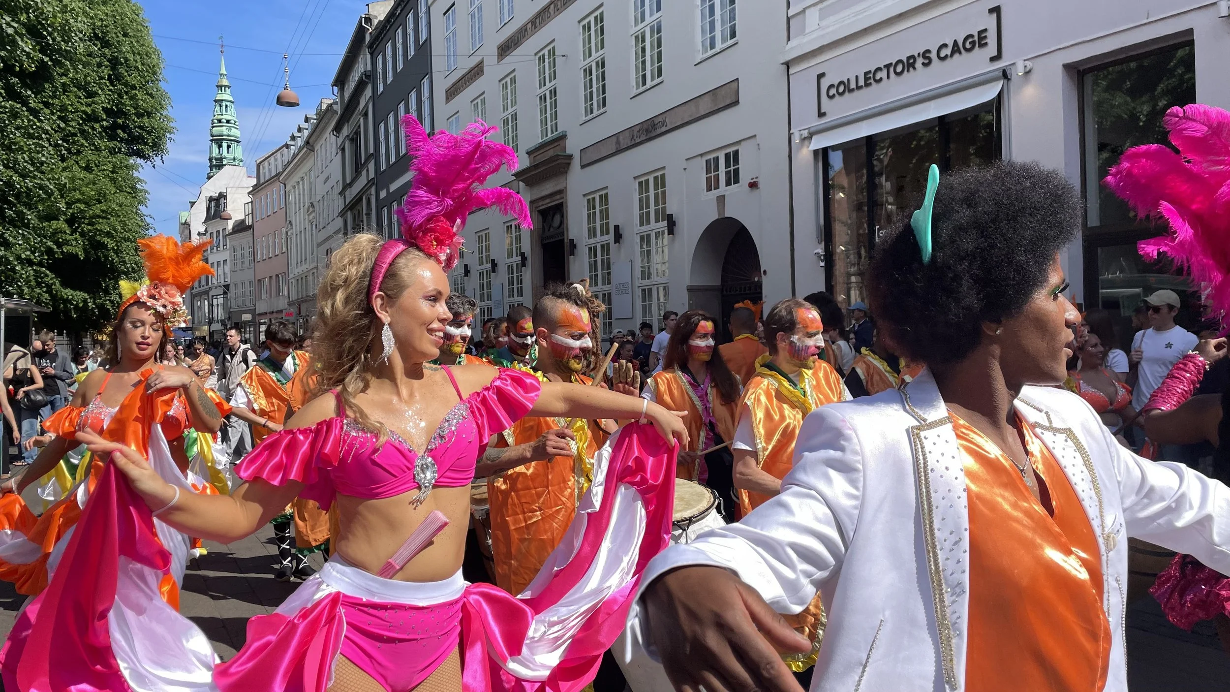 Färgglada karnevalsklädda dansare och musiker som deltar i en parad på en stadsgata under klarblå himmel.