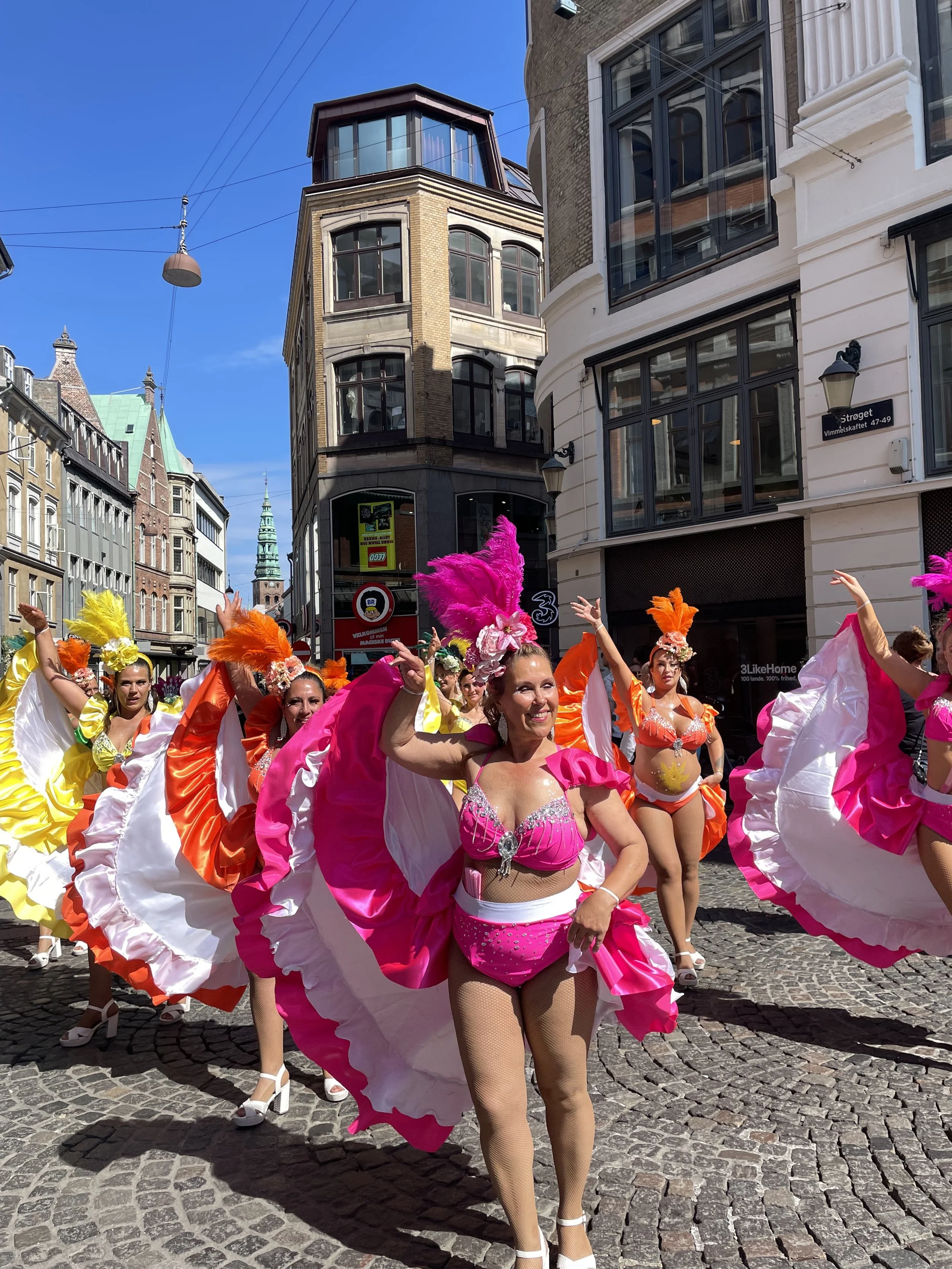 Färgstarka dansare i parade, klädda i rosa, orange, gult och vitt med fjäderhuvor och kjolar, dansar på en gata, bakgrund med gamla byggnader och ett kyrktorn.