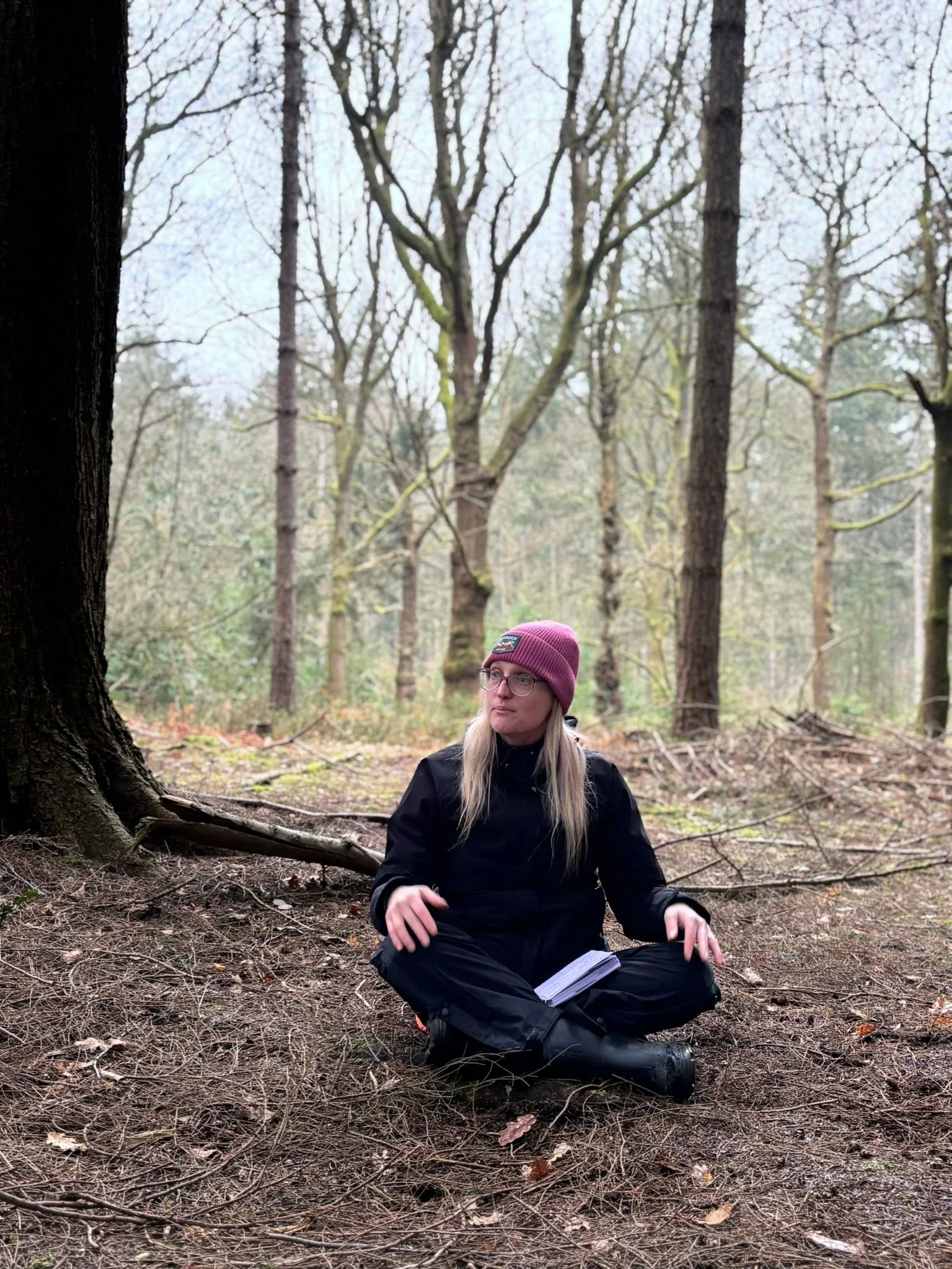 A woman with long blond hair, glasses, and a maroon beanie, sitting on the forest floor with a book in her lap, surrounded by trees in a wooded area.