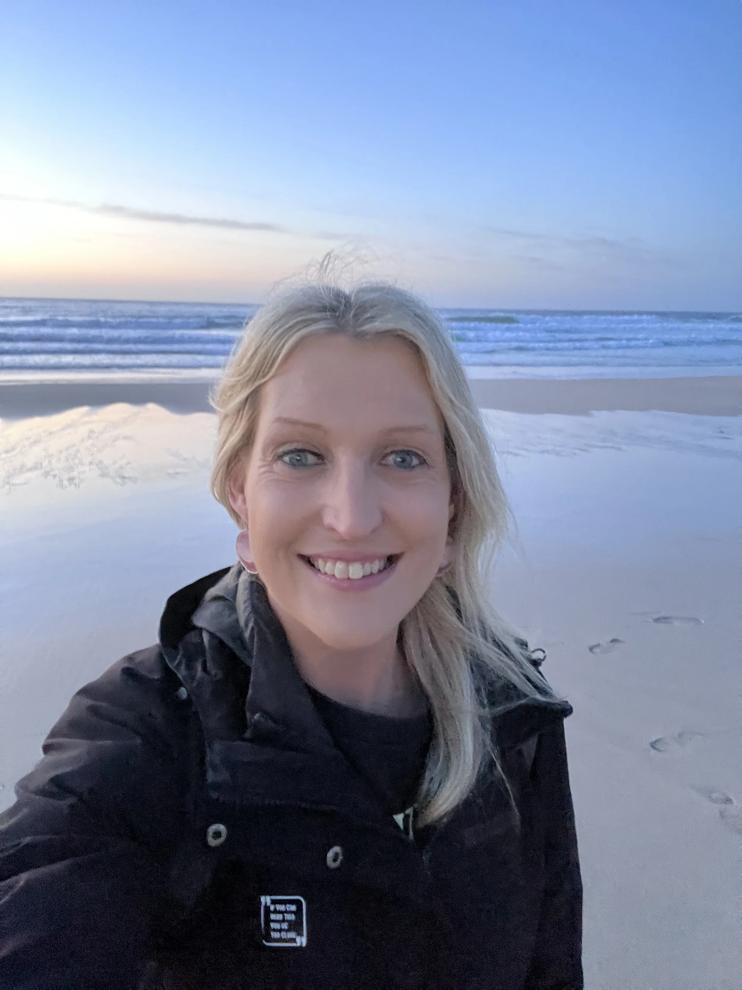 A woman with blonde hair smiling at the camera at the beach during sunset or sunrise, wearing a black jacket.