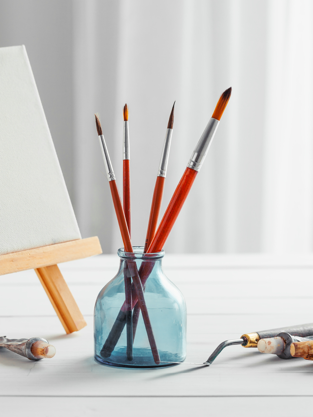 Paintbrushes in a glass jar beside a blank canvas on an easel and a paint roller on a white table.