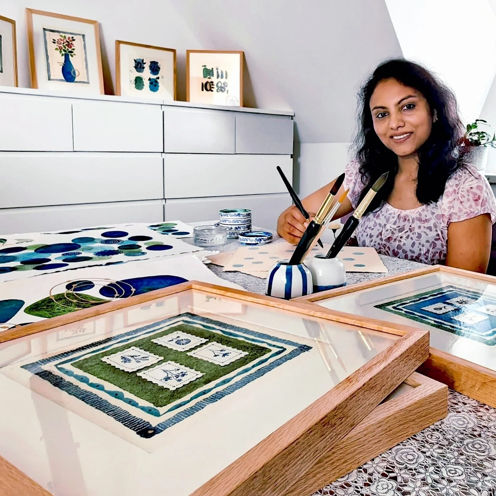 A woman with curly dark hair sitting at a table working on paper craft projects involving blue, green, and white color schemes. The table has several framed fabric or paper pieces, and in the background, there are framed artworks on a white dresser.