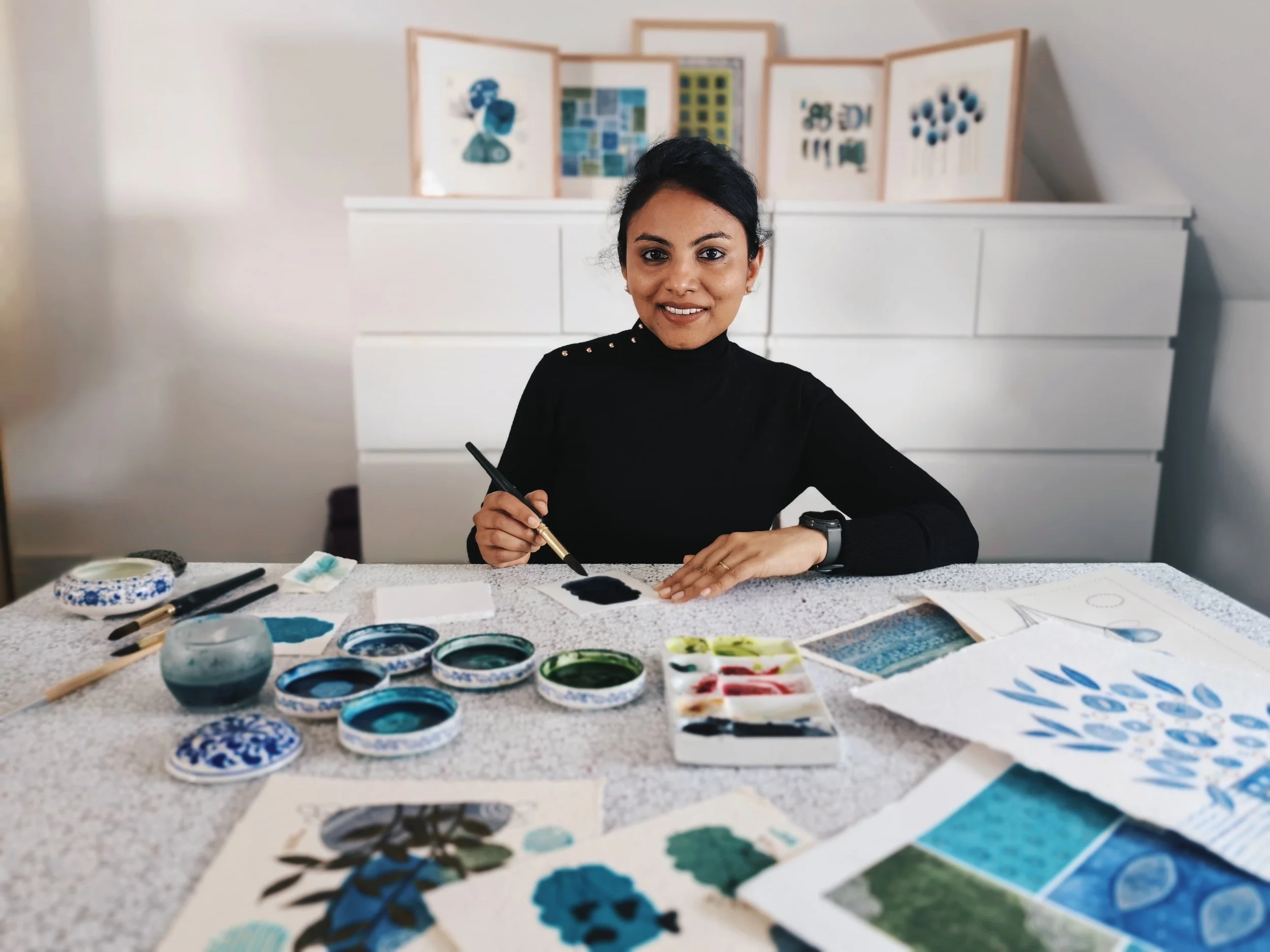 A woman sitting at a table with various paint supplies, including bowls of blue, green, and red paint, paintbrushes, and watercolor paper, creating artwork. In the background, a white cabinet with abstract artwork or posters displayed behind her.