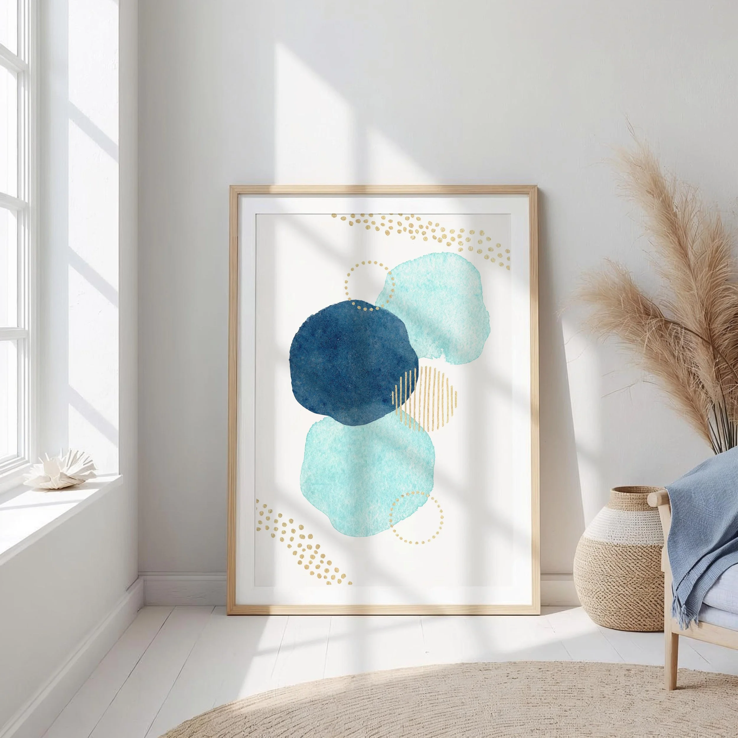 A framed abstract watercolor painting with blue and teal circles, gold dots, and lines, leaning against a white wall near a window with sunlight.