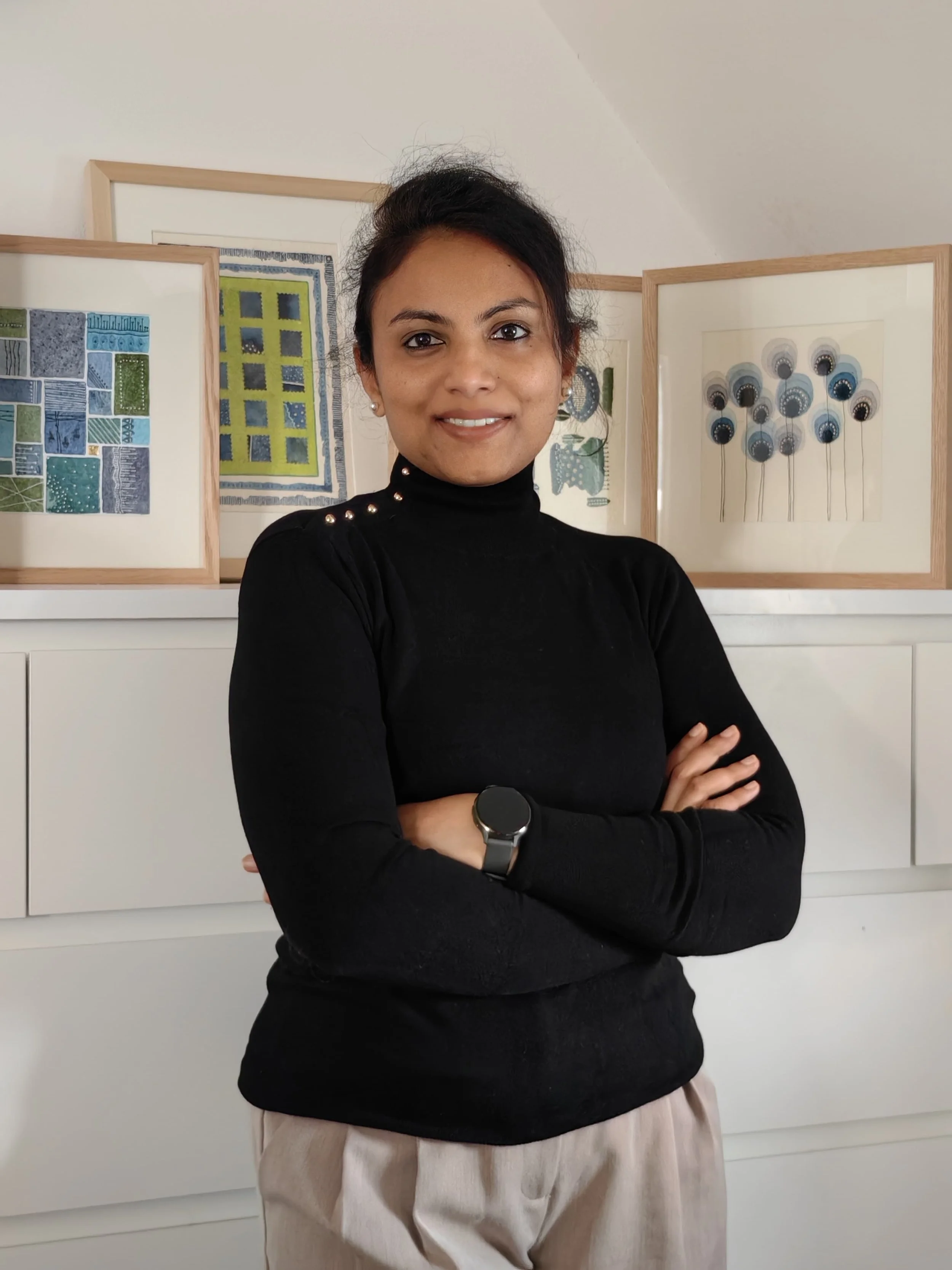 A woman standing indoors with arms crossed, wearing a black turtleneck sweater with pearl embellishments on the shoulders, beige high-waisted pants, silver watch, and small earrings. Background features framed artwork and a white cabinet.