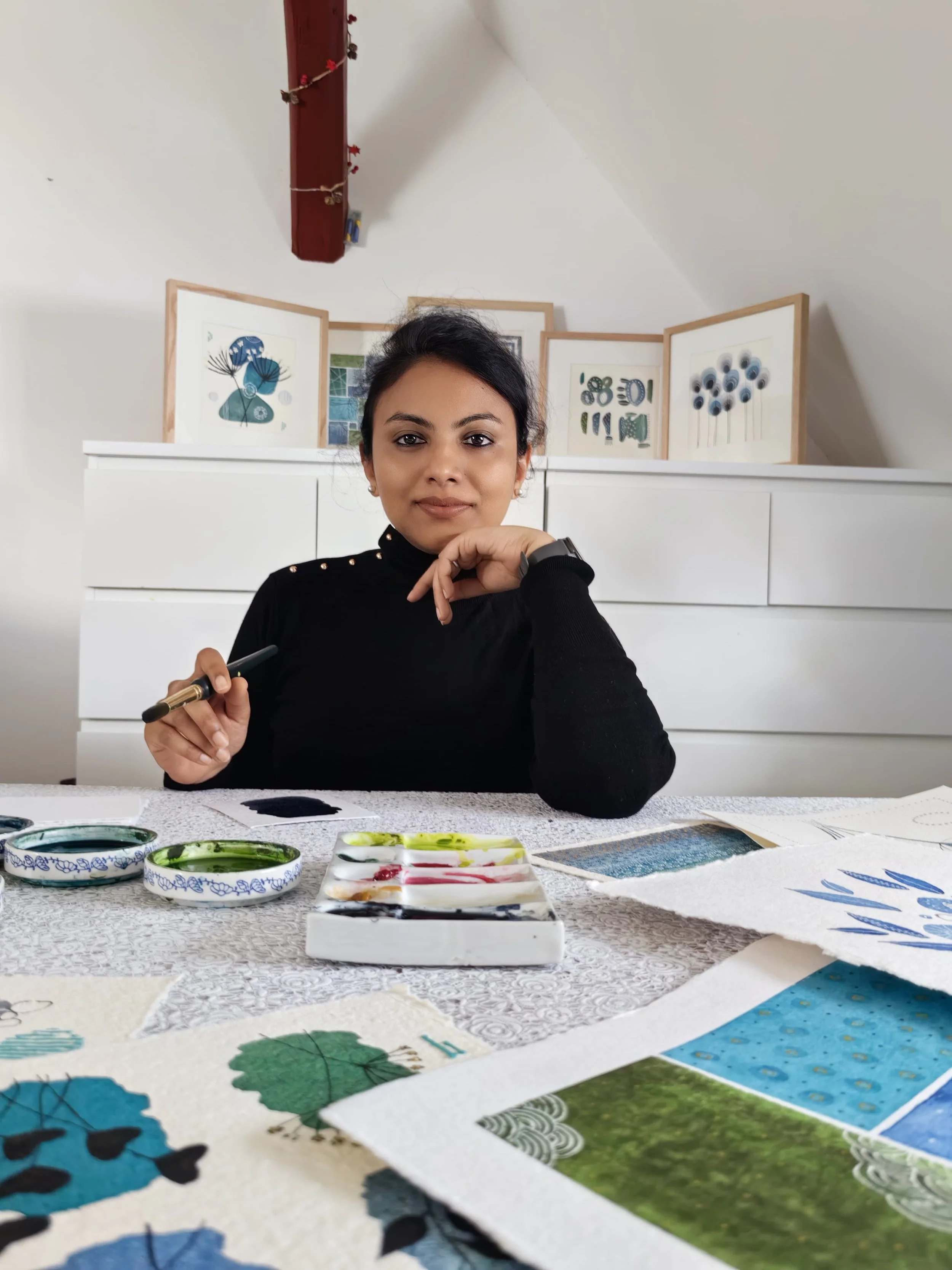 A woman sitting at a table with watercolor paints and watercolor paper, surrounded by her artwork of trees, leaves, and landscapes.