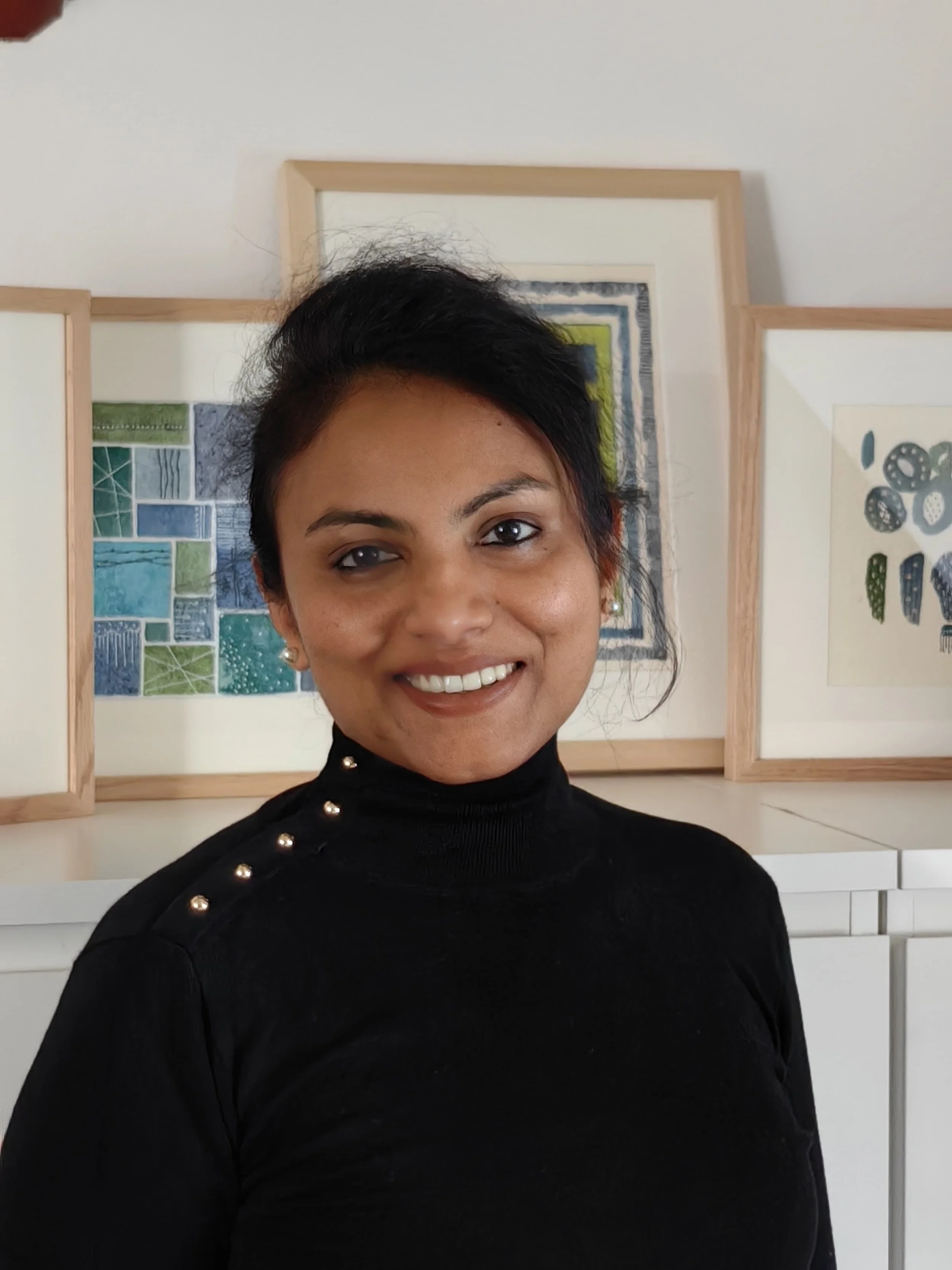 A woman with short black hair smiling in front of artwork on the wall, wearing a black turtleneck sweater with pearl buttons on the shoulder.