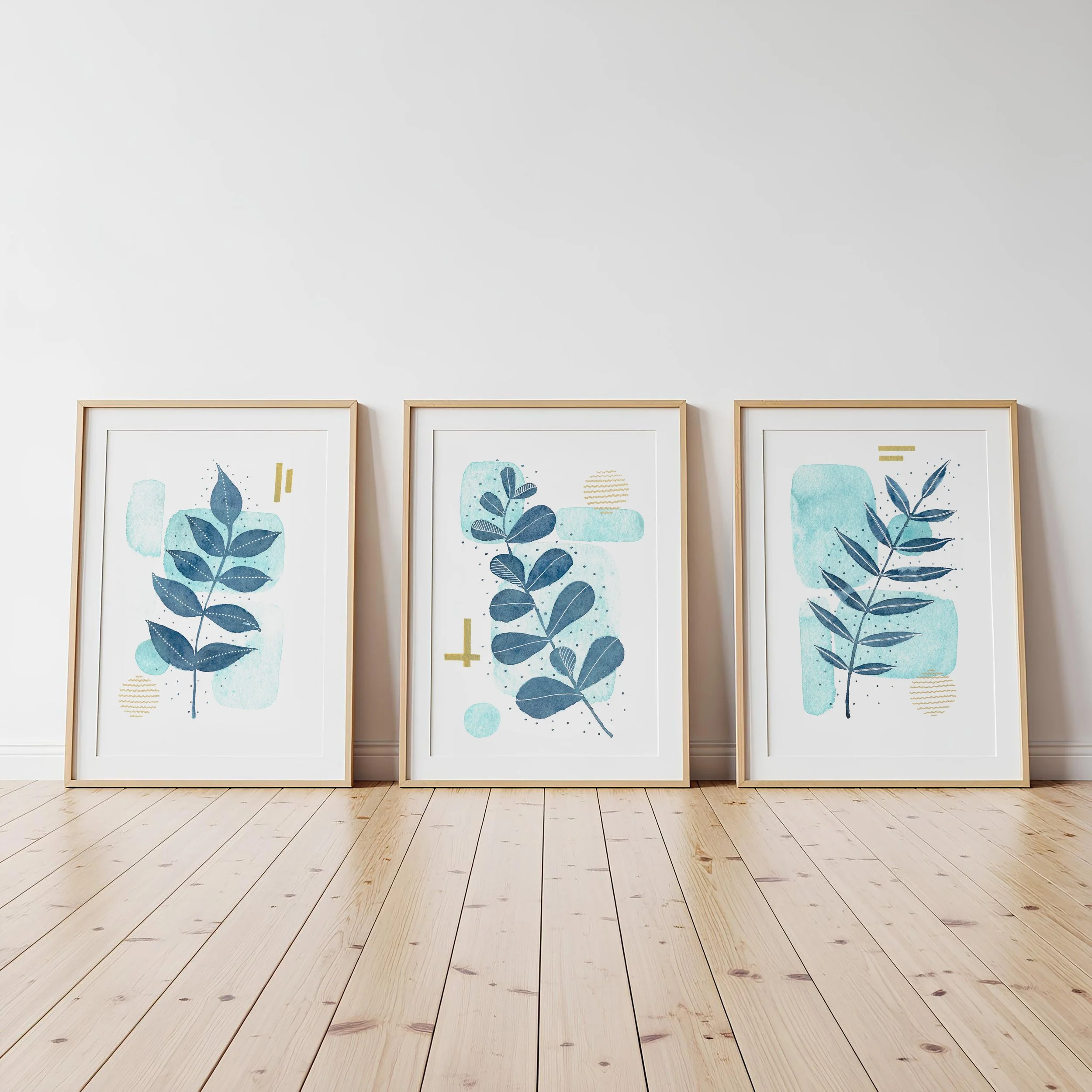 Three framed botanical prints leaning against a plain white wall on a light wooden floor.