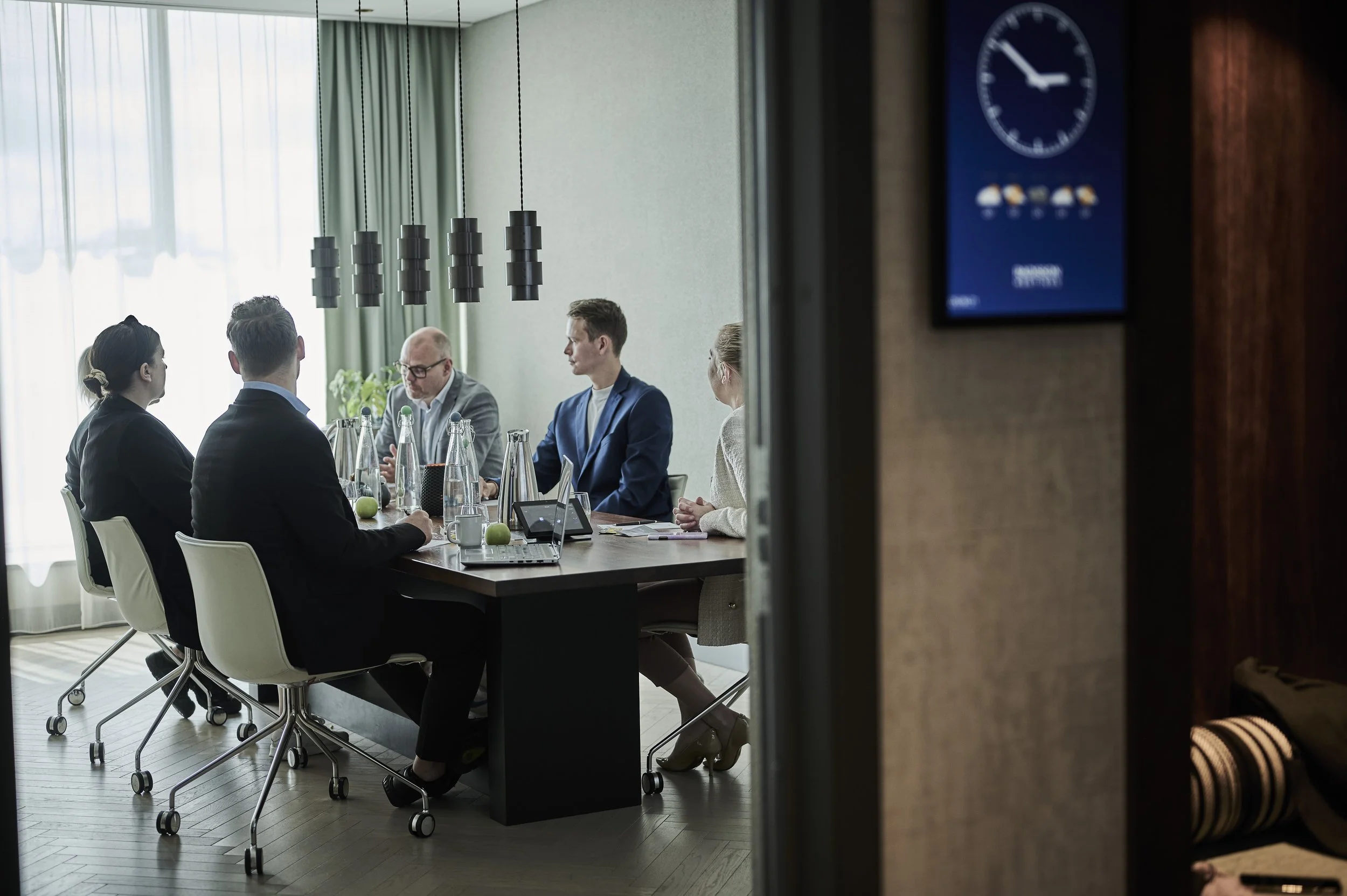 Forskning møter gruppe mennesker i et konferanserom, sett fra siden gjennom en dørkarm, med personer som diskuterer rundt et bord.