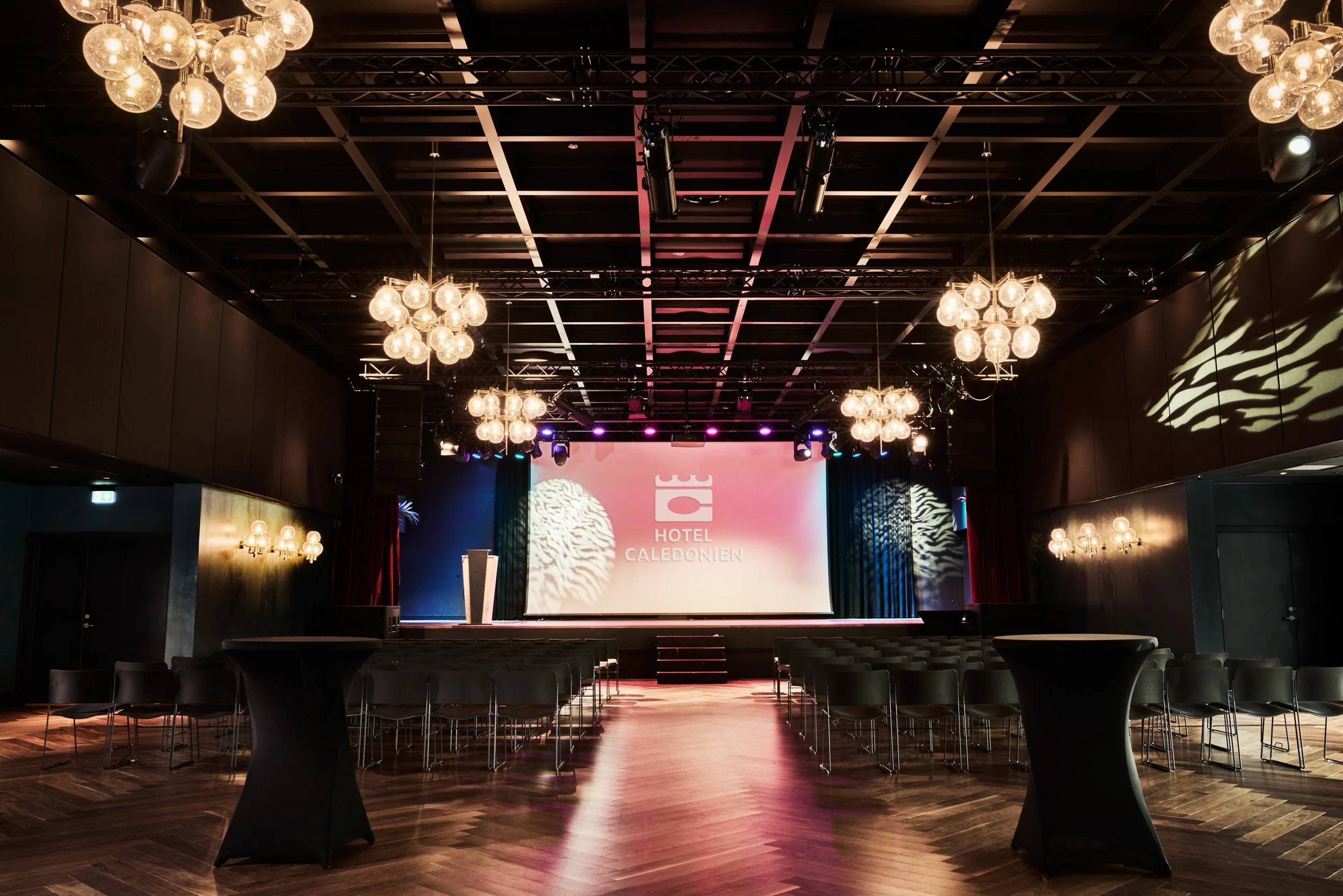 Indendørs konsert- eller konferansesal med flere lysekroner, en stor skjerm med teksten 'Hotel Caledonien', og rader med stoler foran scenen.