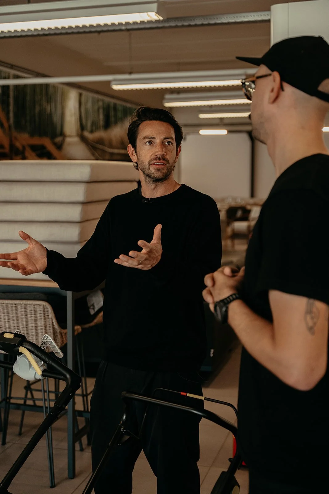 Two men engaged in a conversation indoors, one gesturing with his hands, with furniture and stacked mattresses in the background.