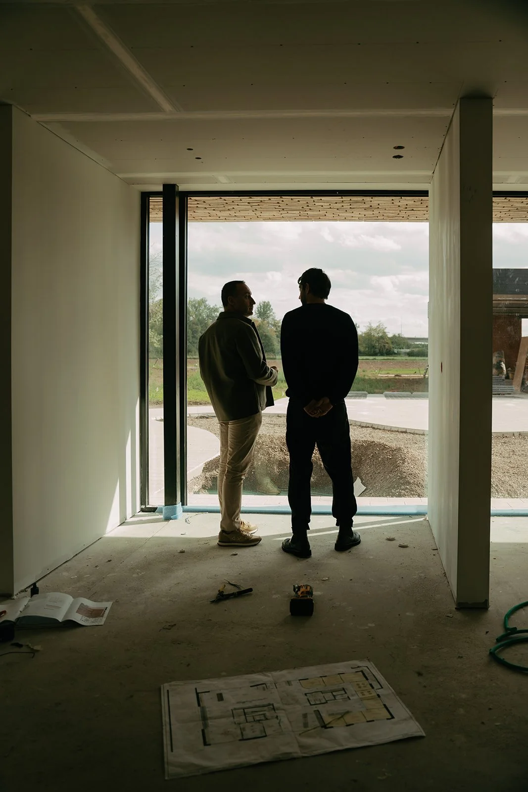 Two men in conversation inside an unfinished building, looking out at a construction site with green fields and partly cloudy sky in the background.