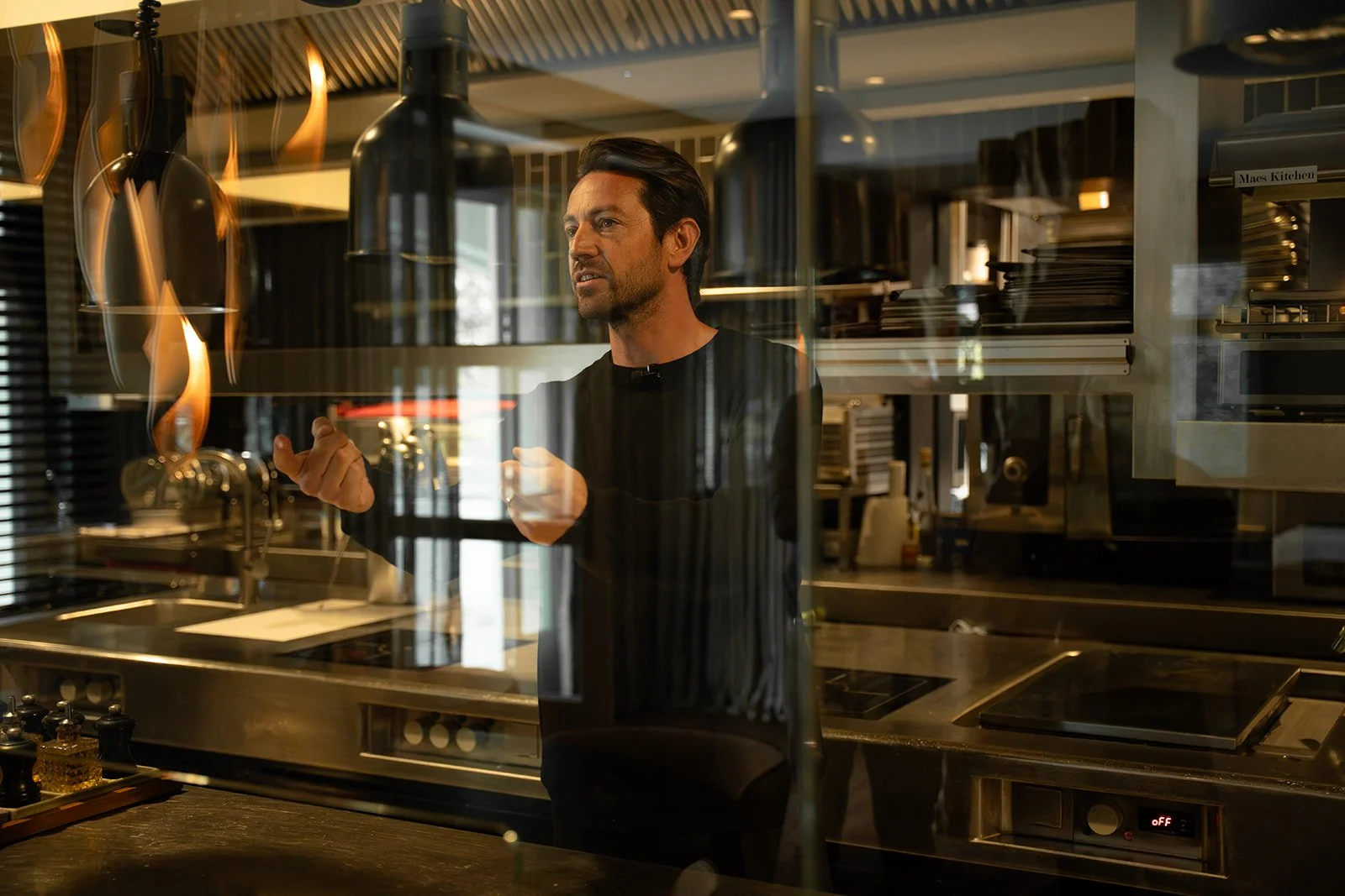 Man inside a restaurant kitchen, seen through a glass window, with flames and kitchen equipment in background.